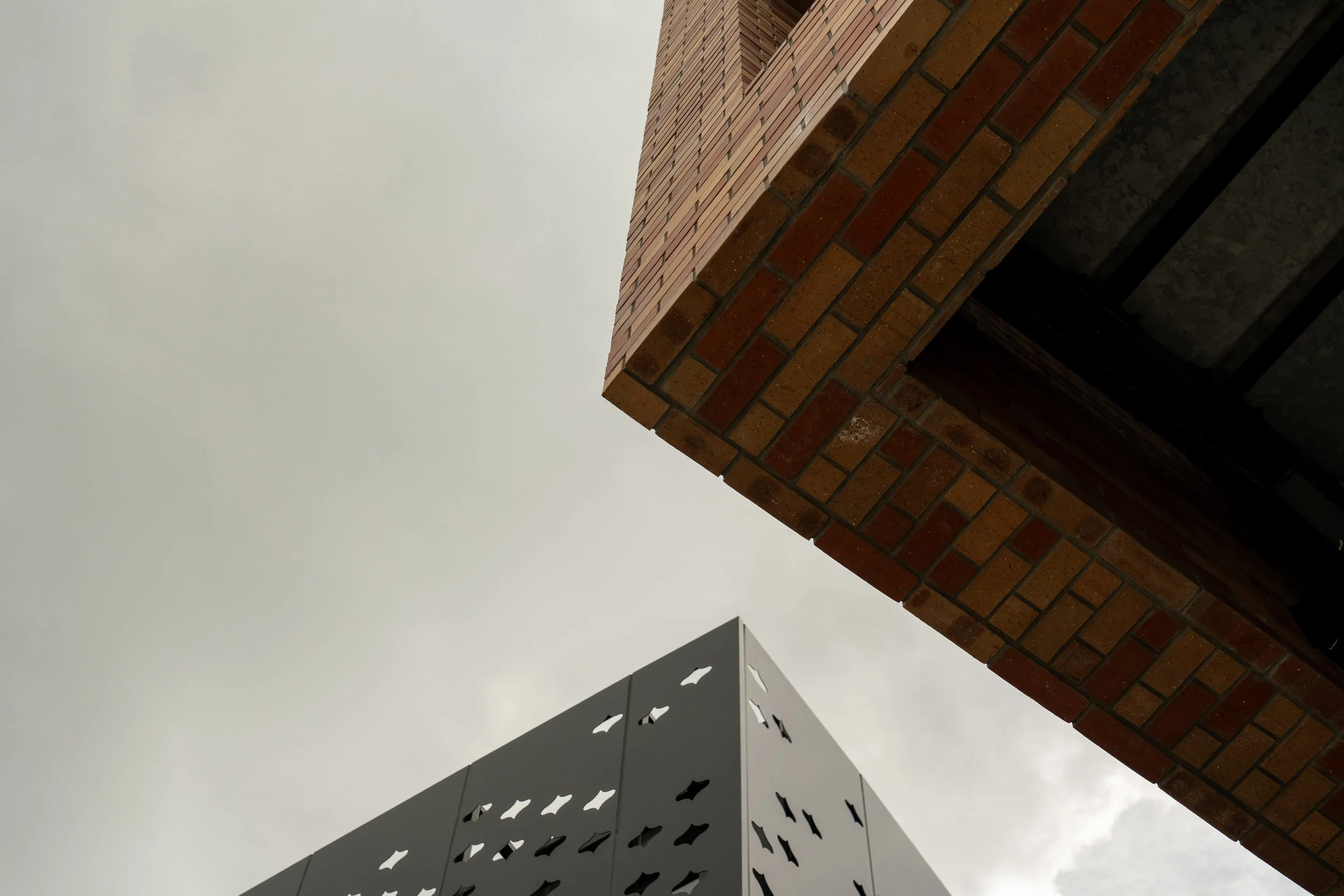 Low-angle view of the sky with the corners of two modern buildings, one with a concrete face with window cutouts and the other with a brick facade, pointing towards an overcast sky.