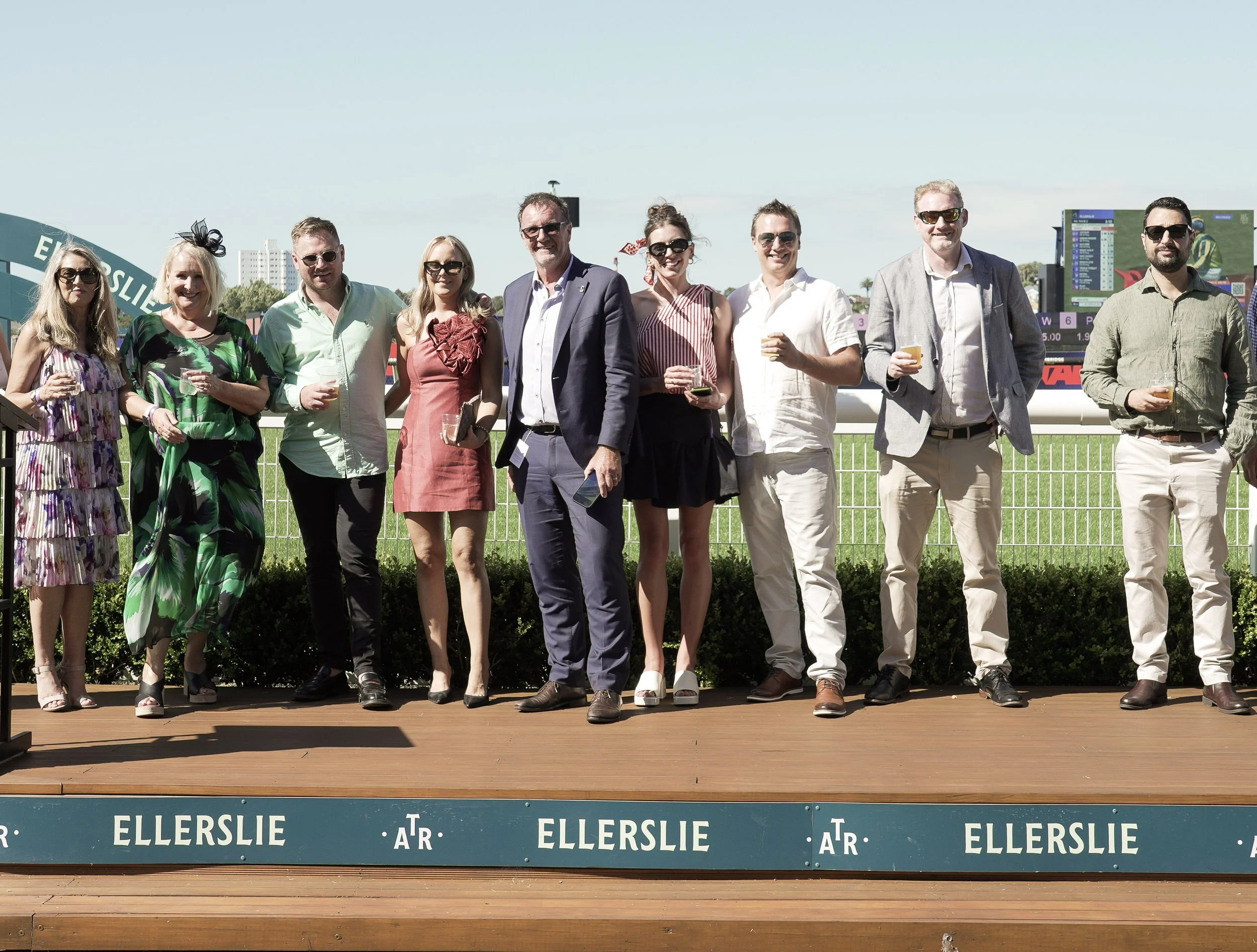 A group of ten people standing on a wooden platform at a horse racing track, with drinks in hand, posing for a photo. The background shows a digital screen and a grassy racecourse, with clear weather.