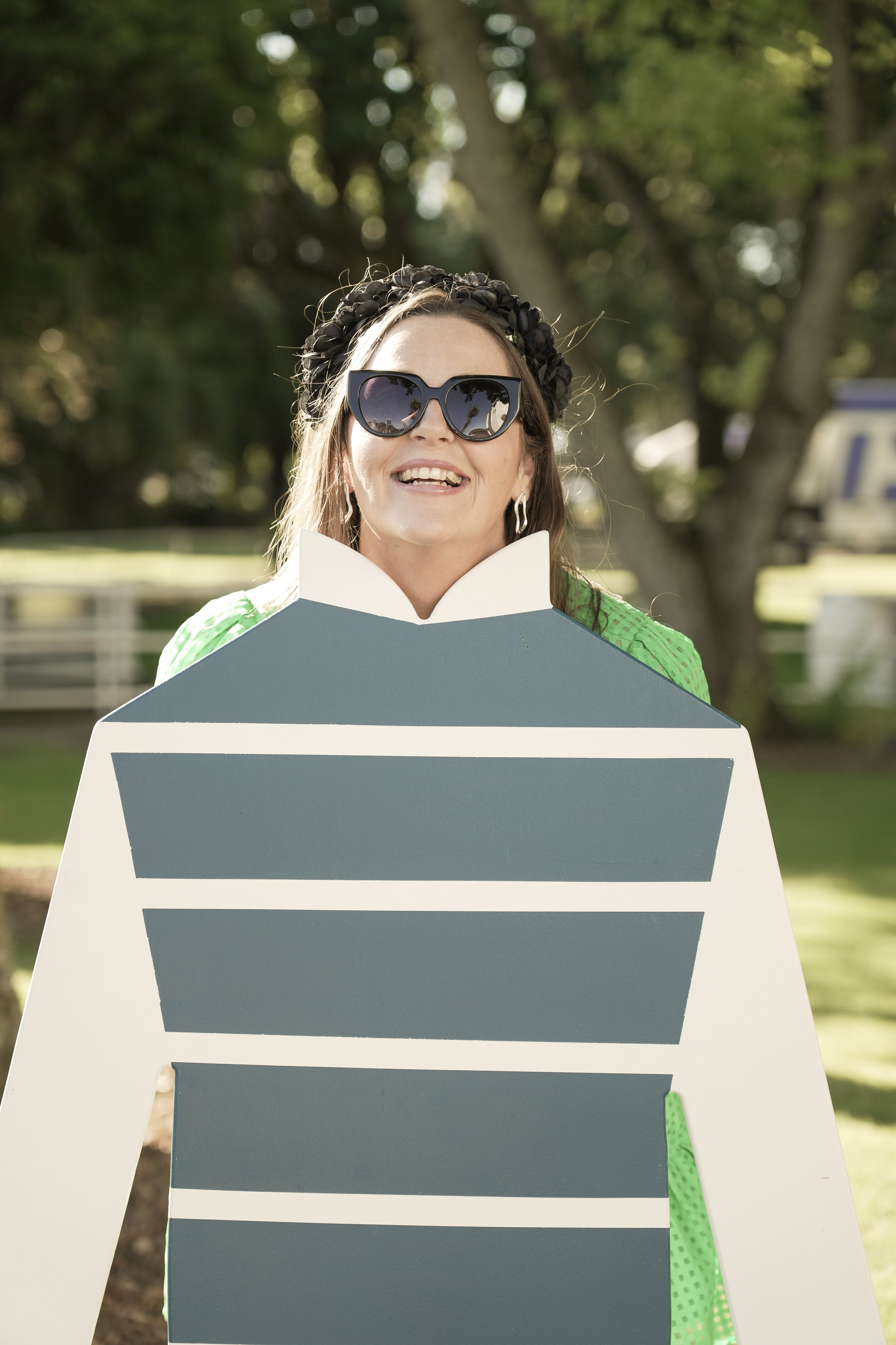 Woman wearing sunglasses and a black headband, smiling behind a large, colorful, striped board outdoors.
