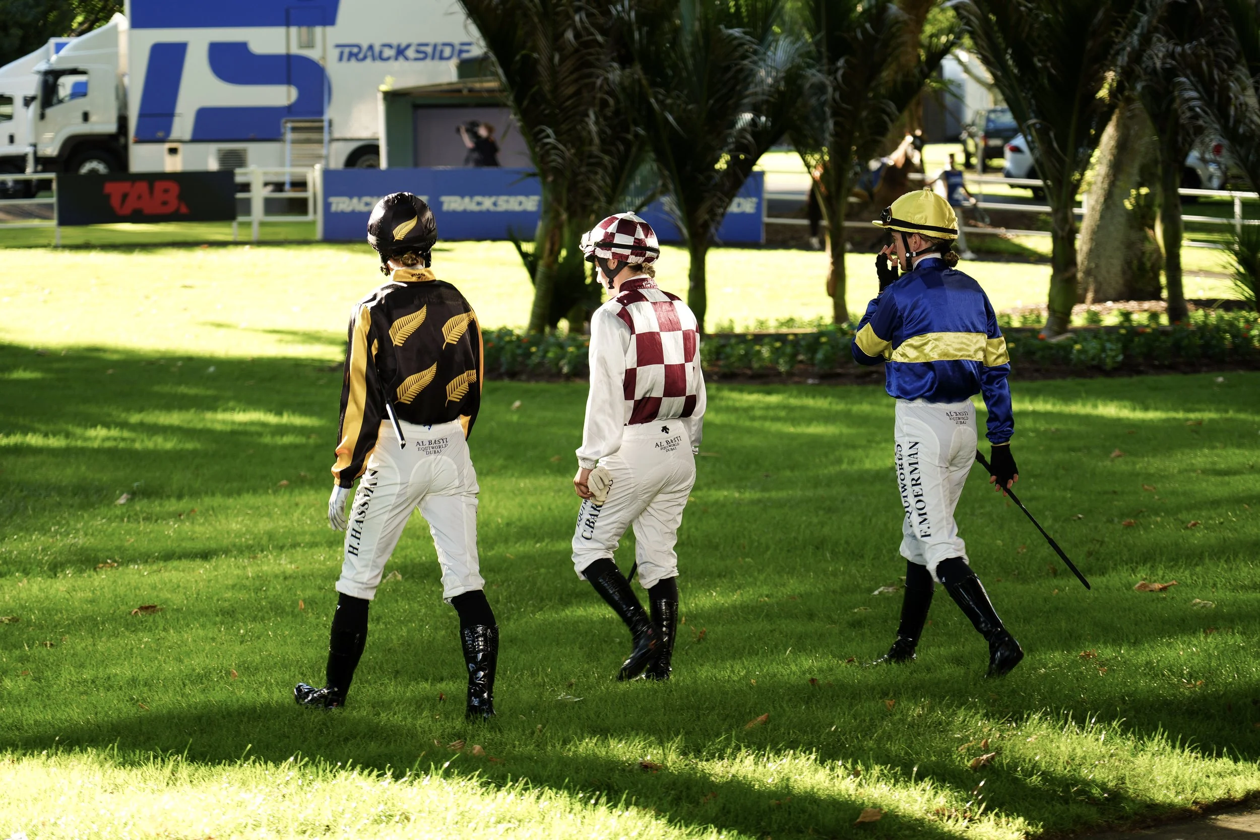 Three jockeys dressed in colorful racing silks and white riding pants walk on a grassy area at a racetrack, with a backdrop of trees, racetrack signs, and trailers.