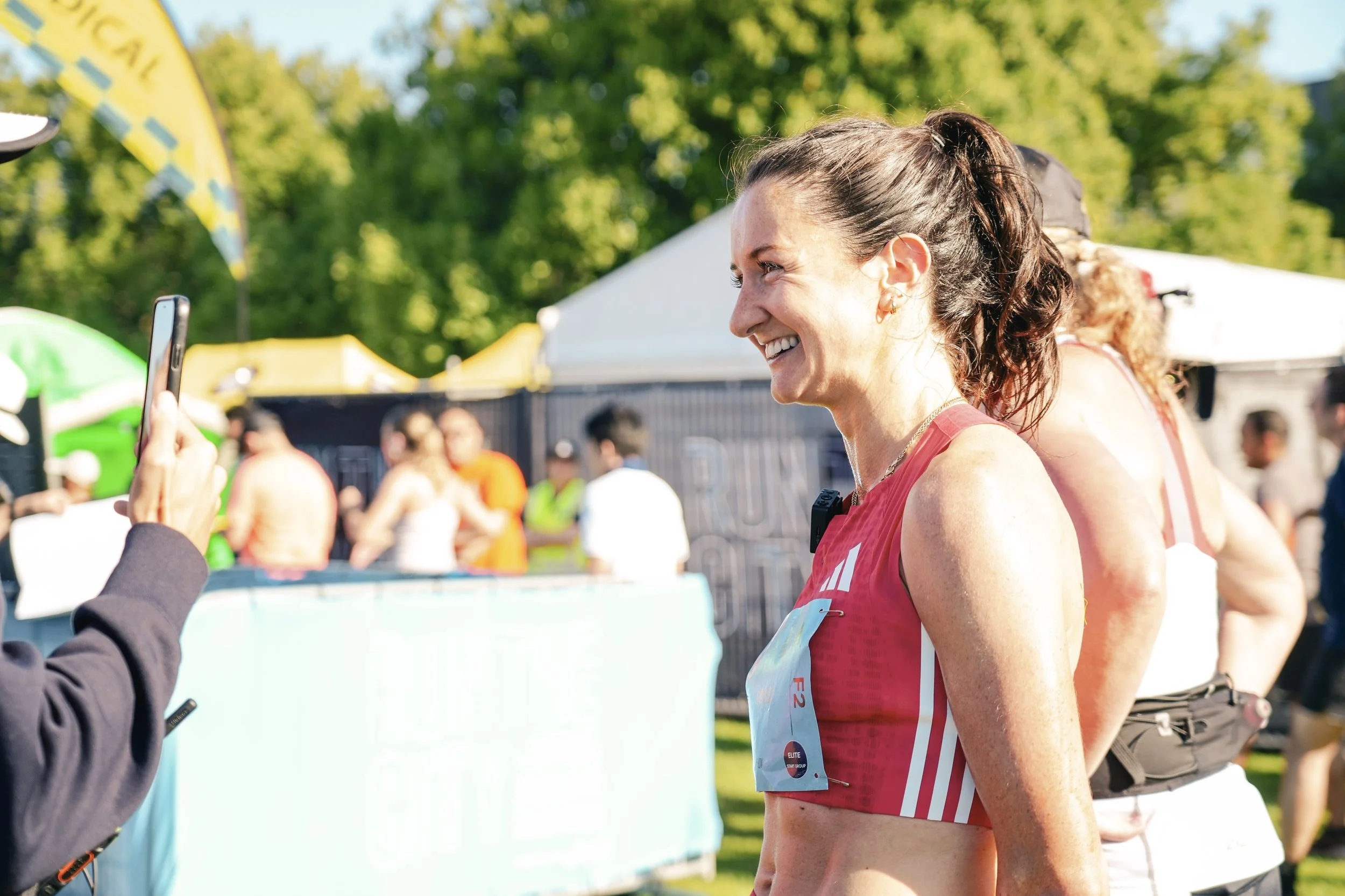 Smiling woman in athletic attire being photographed at an outdoor event with people, tents, and green trees in the background.