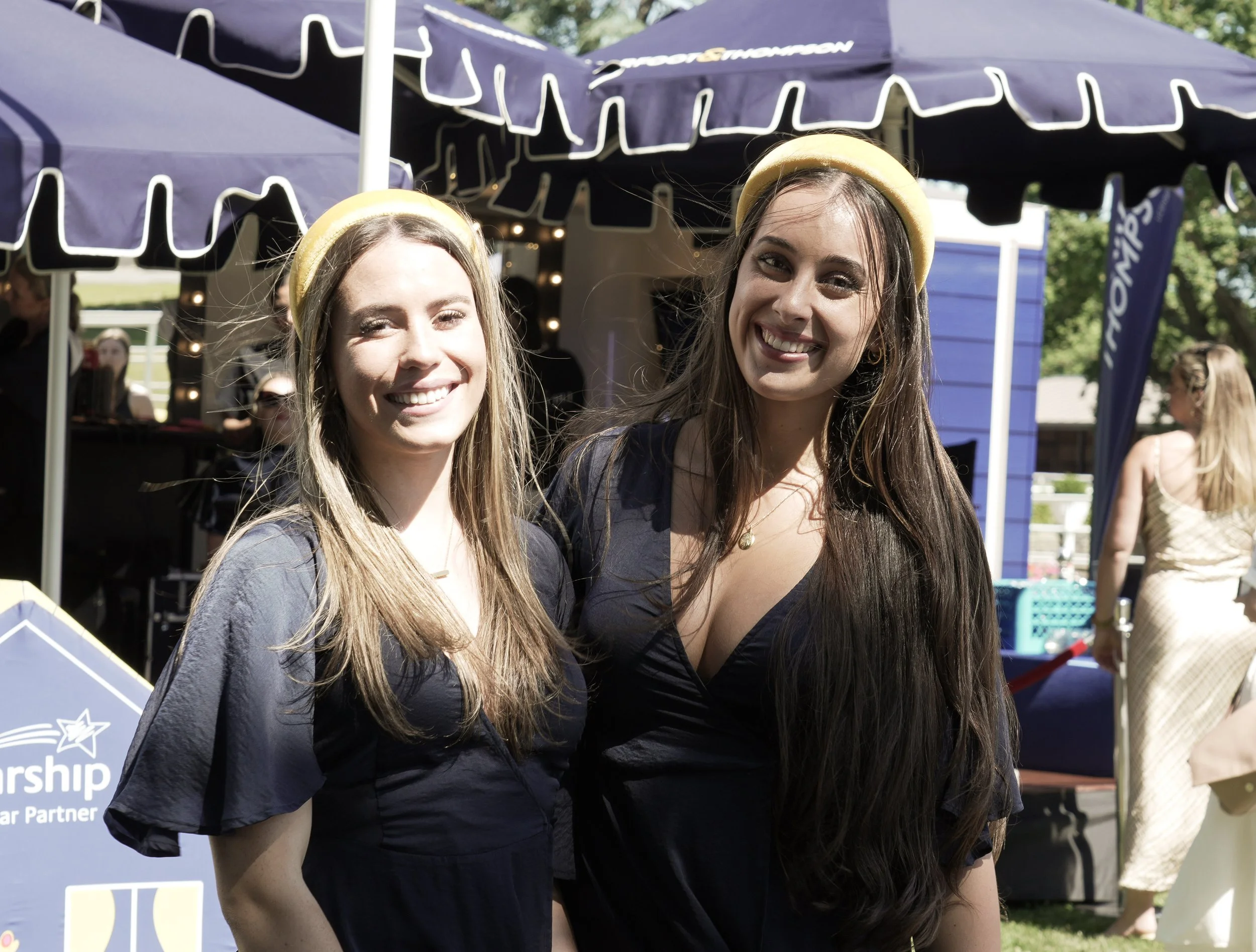 Two smiling women with long hair wearing yellow headbands and black dresses standing outdoors at an event, with booths and people in the background.