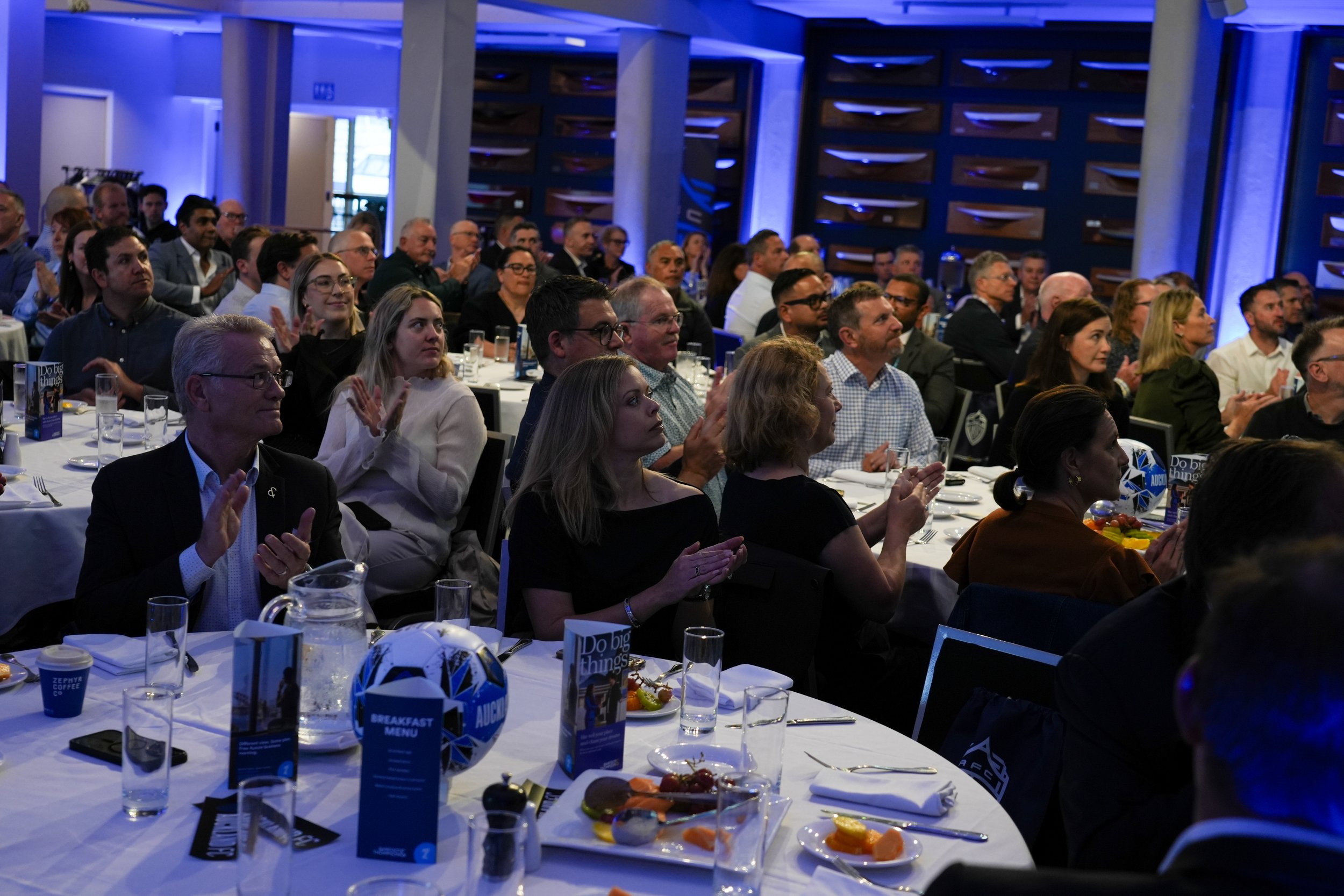 attendees at a conference or event sitting at round tables, clapping and listening to a speaker or presentation in a dimly lit room with blue lighting.