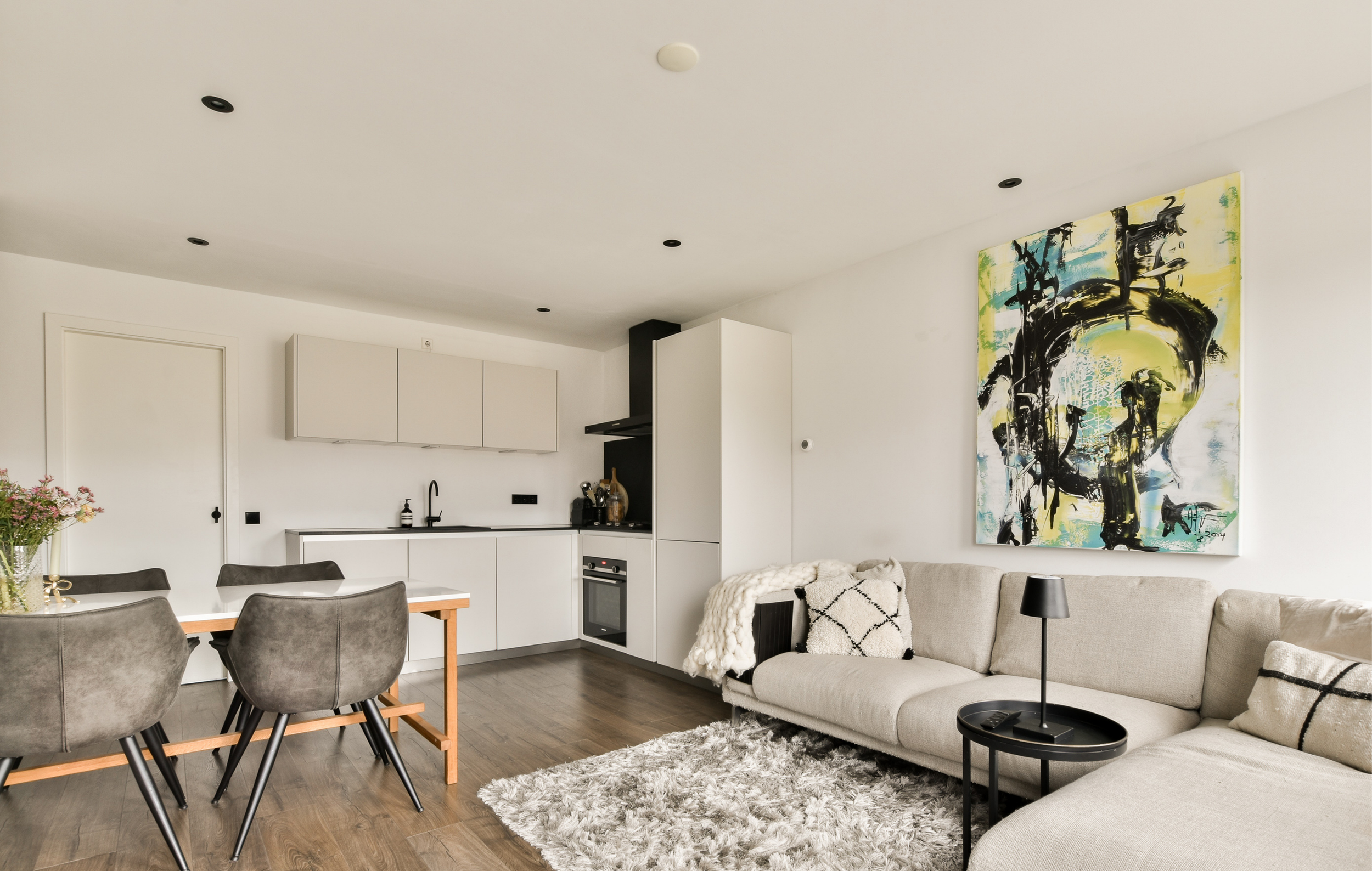 Modern living room and kitchen with white walls, beige sofa with throw pillows, black side table with black lamp, abstract artwork on wall, dining table with gray chairs, wooden flooring.