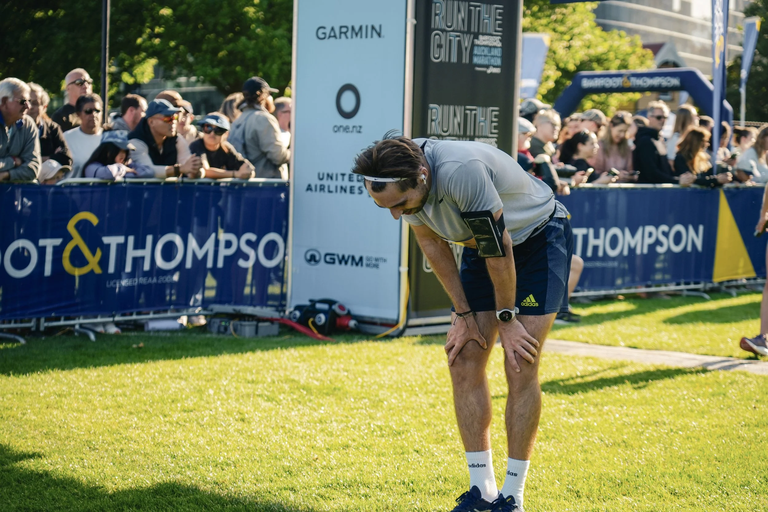 Runner in gray shirt and navy shorts bends over, holding his knee, during a race event, with spectators behind blue barricades and event signage.