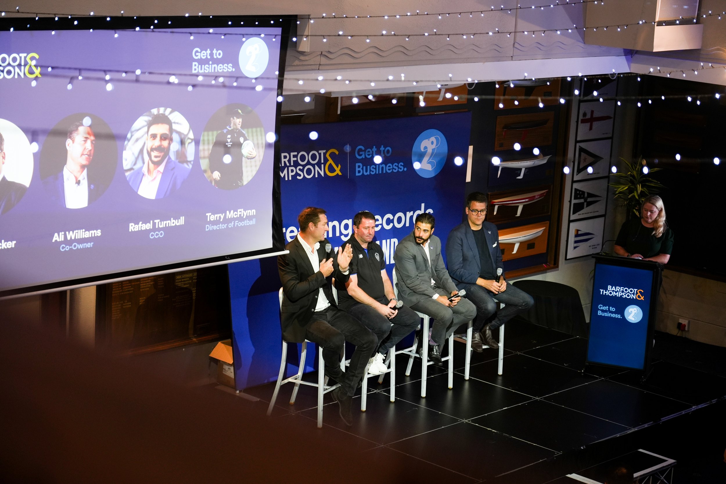 A panel discussion at a conference featuring four men sitting on stools on stage, with a large screen displaying speakers' pictures and names to their left. The screen shows three speakers: Ali Williams, Rafael Turnbull, and Terry McFynn. A woman sta