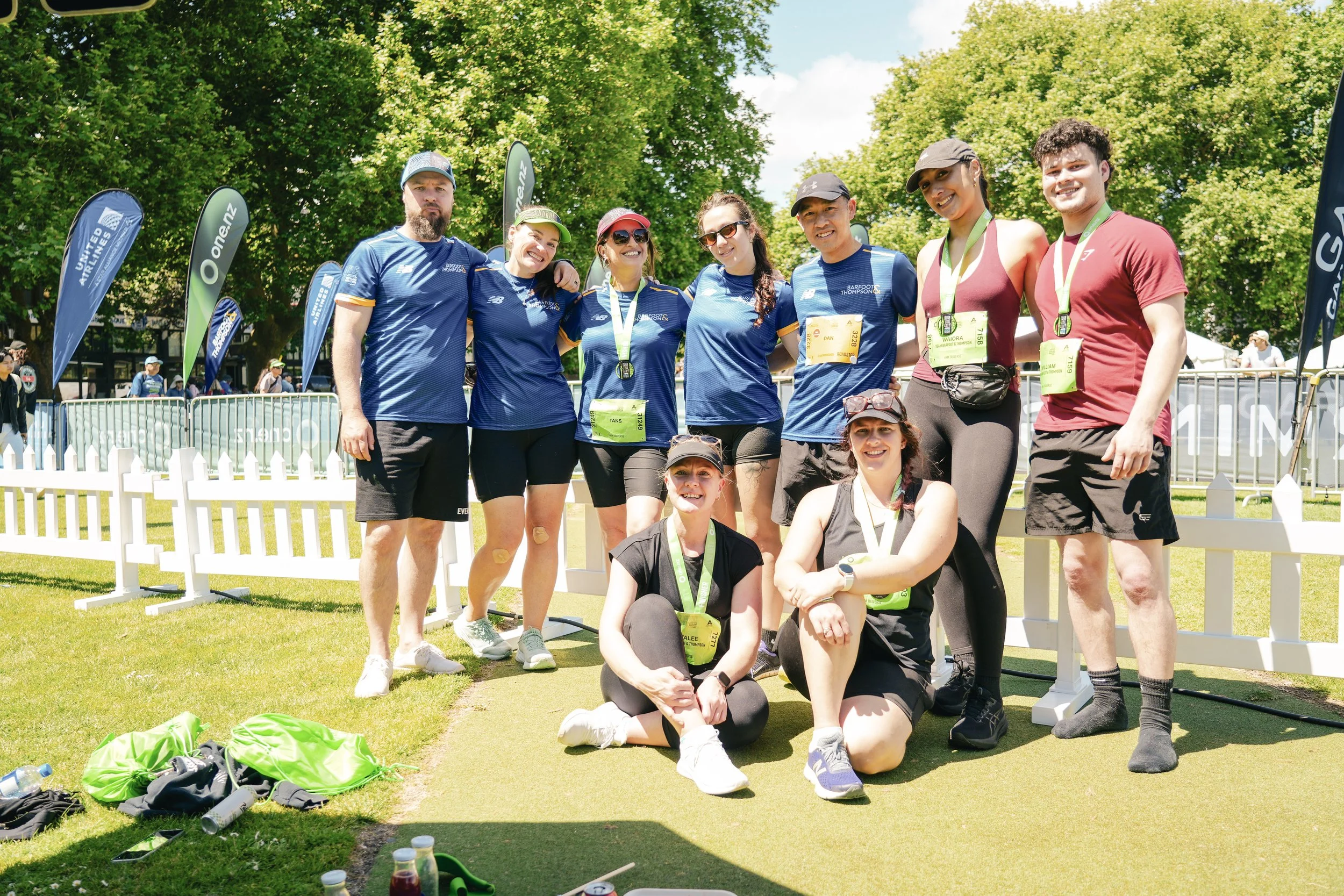 Group of nine runners, some wearing medals, posing for a photo outdoors on a sunny day after a race, with green trees and race flags in the background.