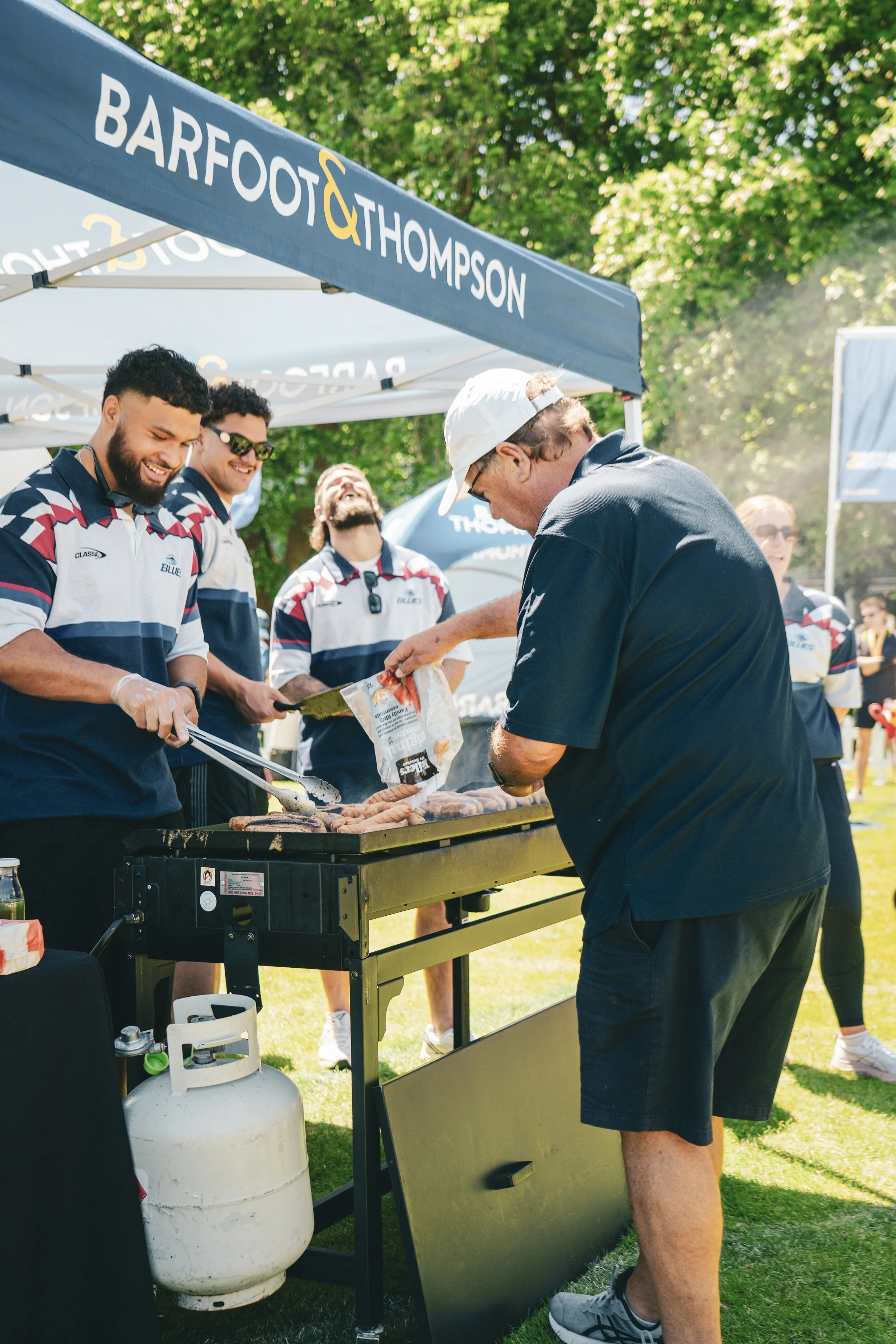 A group of men cooking and grilling sausages at an outdoor event under a blue canopy tent with the text 'Barfoot & Thompson'. They are diverse, wearing similar striped shirts, smiling, and preparing food on a grill with a propane tank underneath. Tre