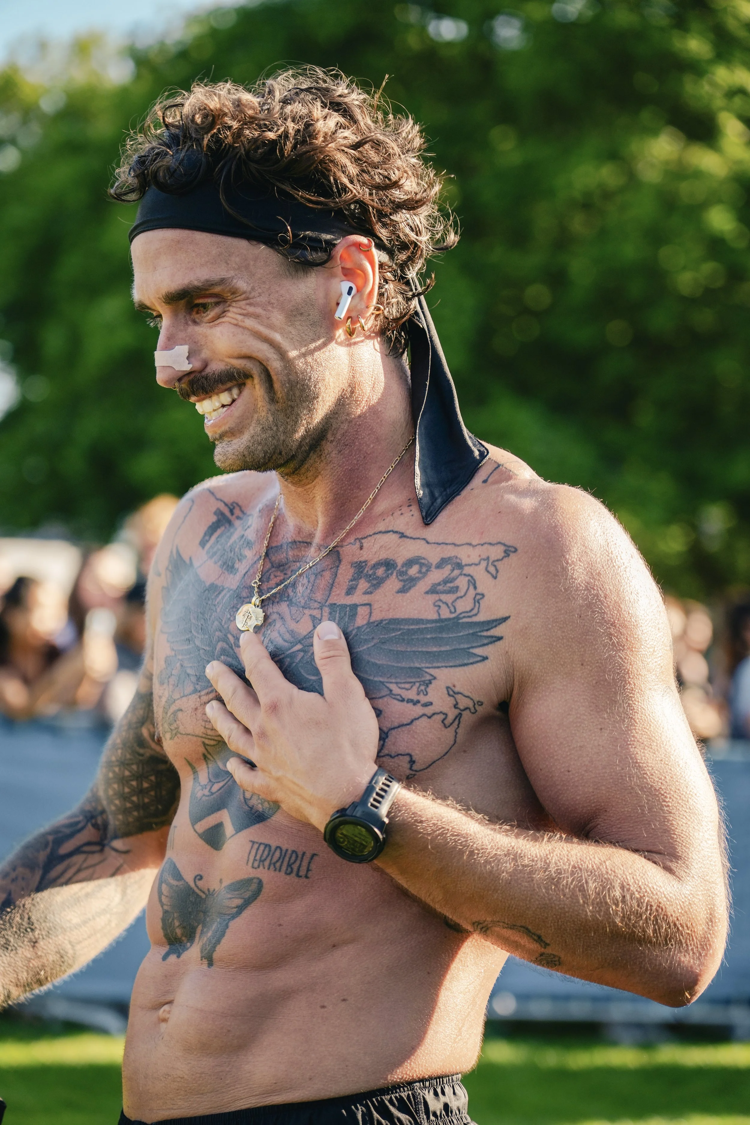 A smiling man with tattoos, earphones, and a black headband, crossing his hand over his heart, outdoors on a sunny day.