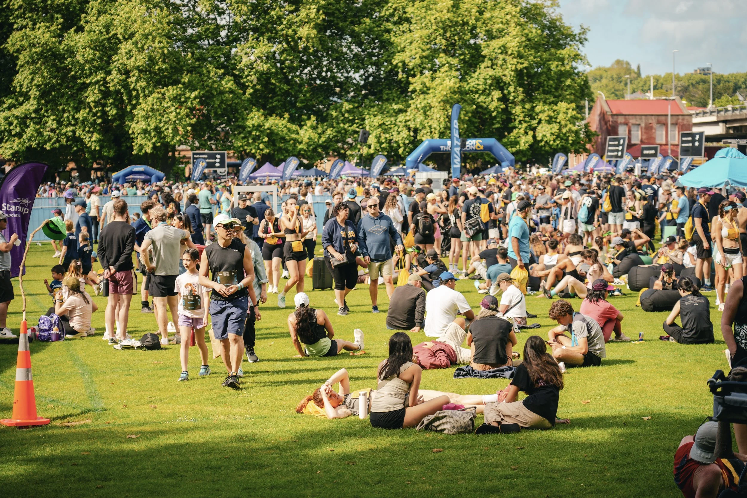 A large crowd of people gathered outdoors on a grassy field, some sitting and some walking, with tents and colorful banners in the background under a sunny sky.