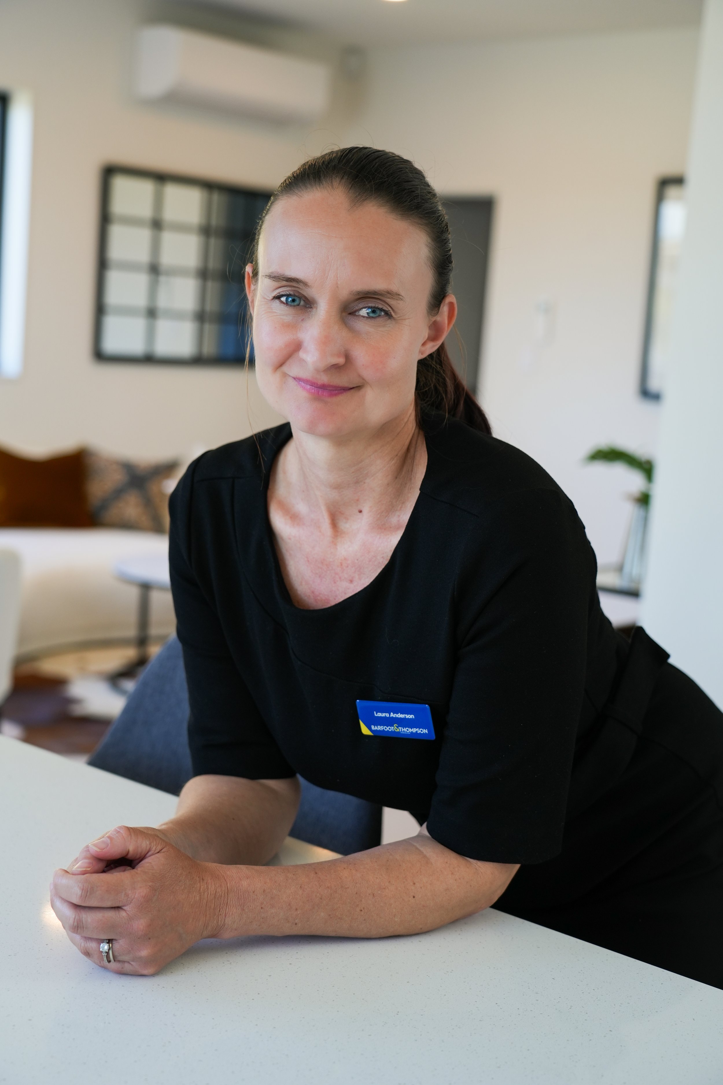 A woman with light skin, blue eyes, and dark brown hair tied back, wearing a black shirt with a name tag, sitting at a white table in a bright, modern living room with framed pictures on the wall and a beige sofa in the background.