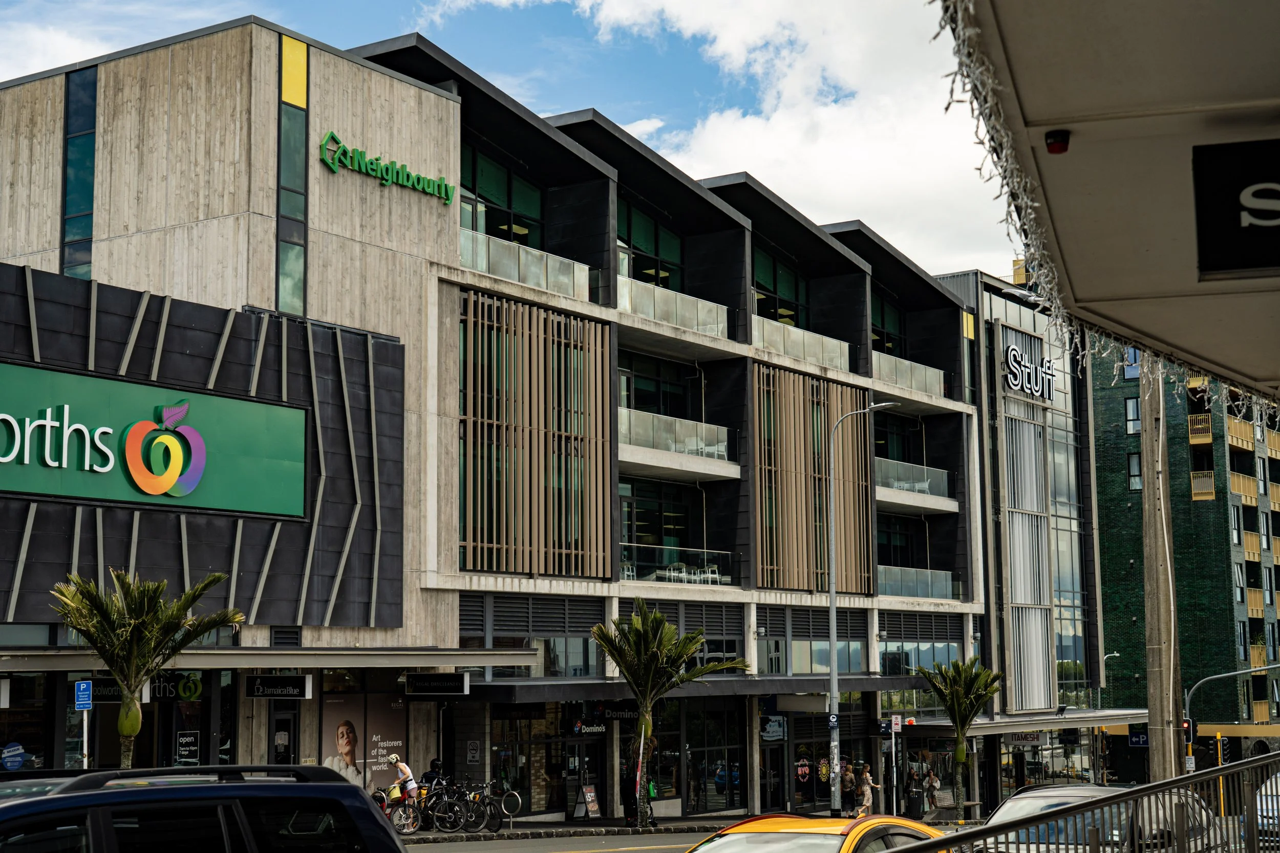 Modern multi-story shopping mall with stores like Woolworths, STUFF, and Domino's Pizza on the ground level. The building has wooden and black paneling, glass balconies, and vertical wooden slats. There are palm trees in front, bicycles parked outsid