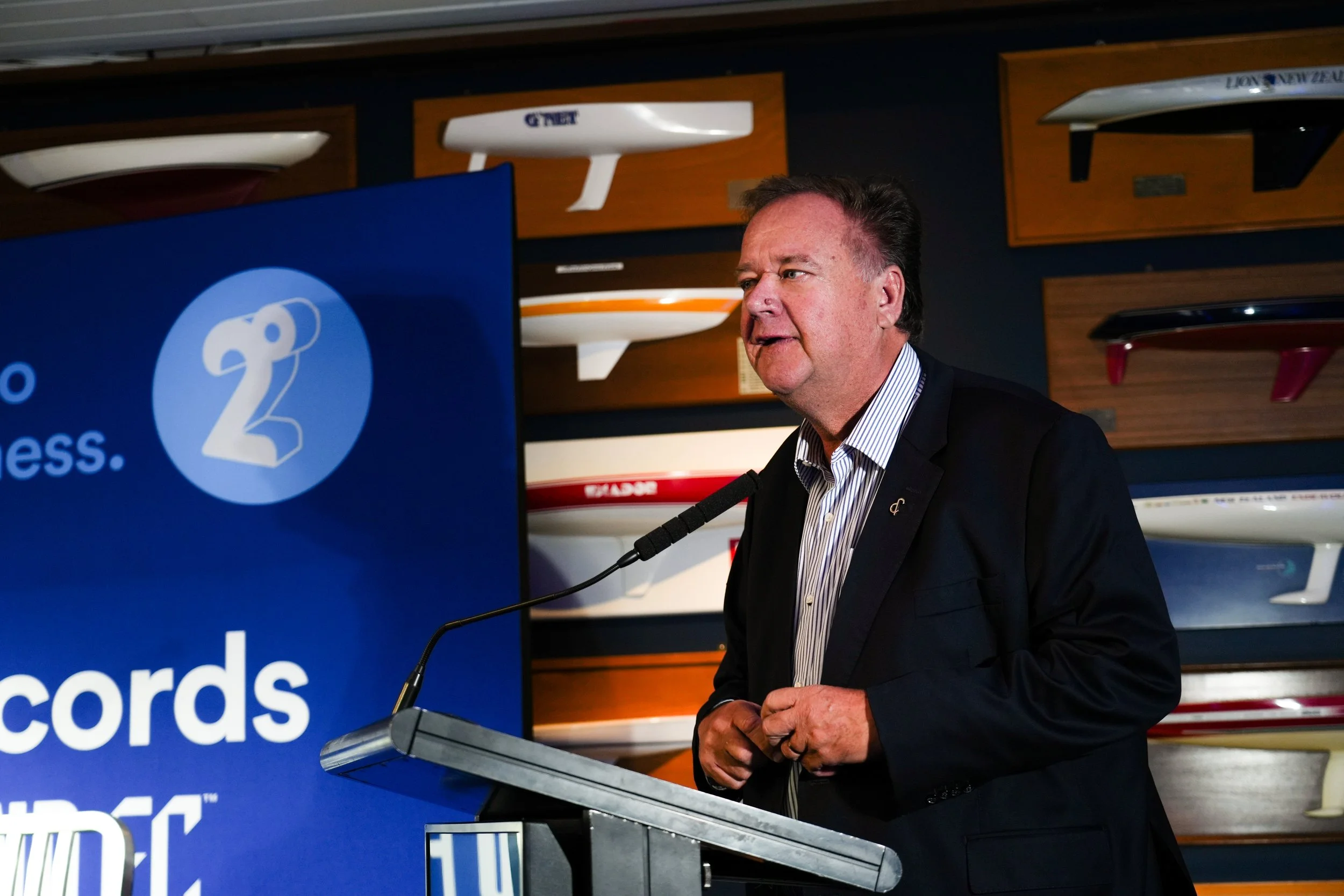 Man in a suit speaking at a podium with a large screen displaying the text 'records' and a logo with the number 2, with surfboards mounted on the wall behind him.