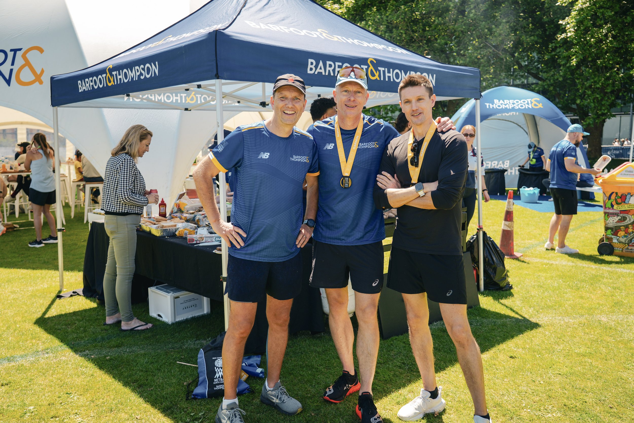 Three men wearing blue and black athletic clothing with medals around their necks posing in front of a blue tent at an outdoor event on a sunny day. Other people and event tents are visible in the background.