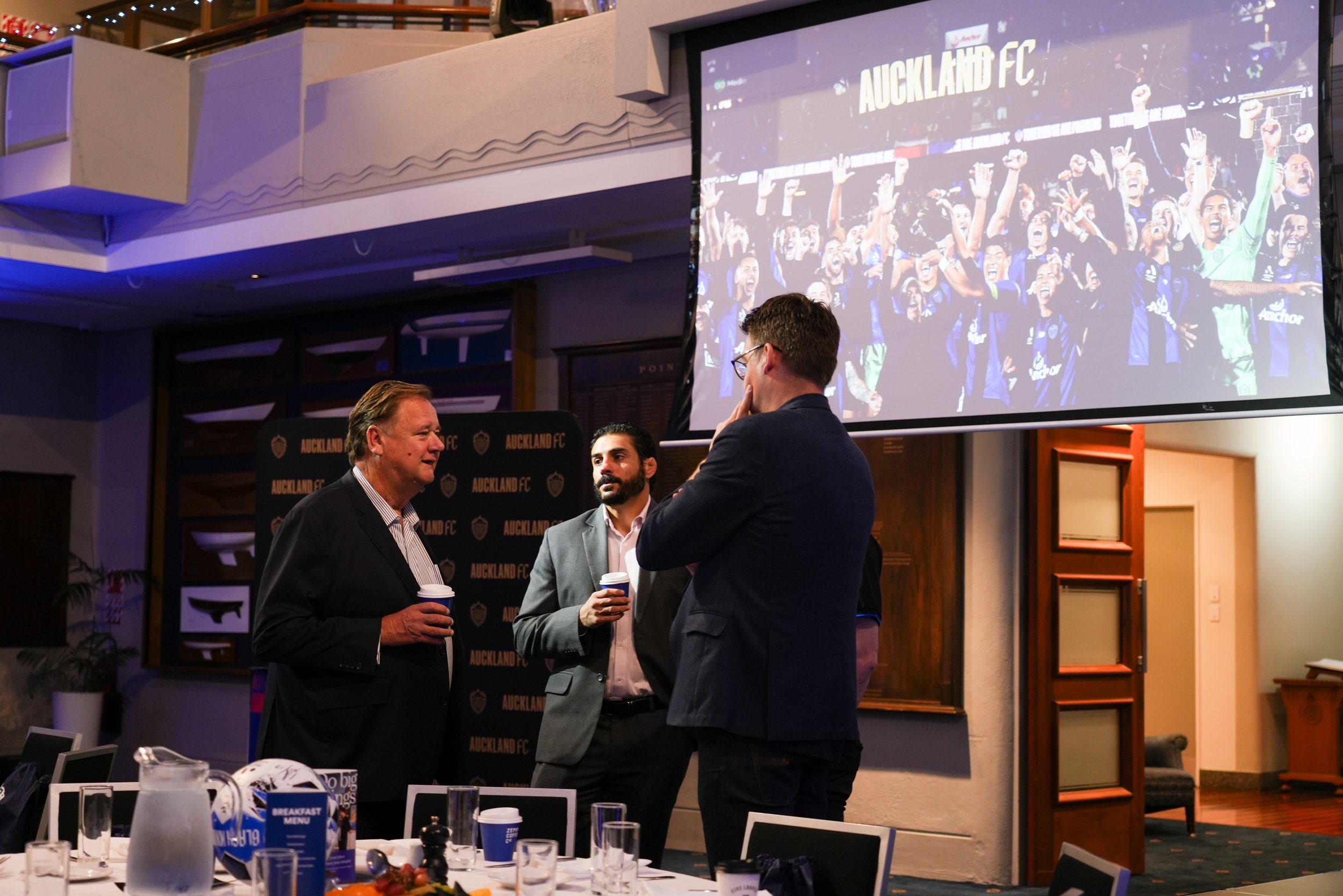 Three men in formal attire having a conversation in front of a large screen and a backdrop with Auckland FC branding, with a table set with glasses and a pitcher in the foreground.