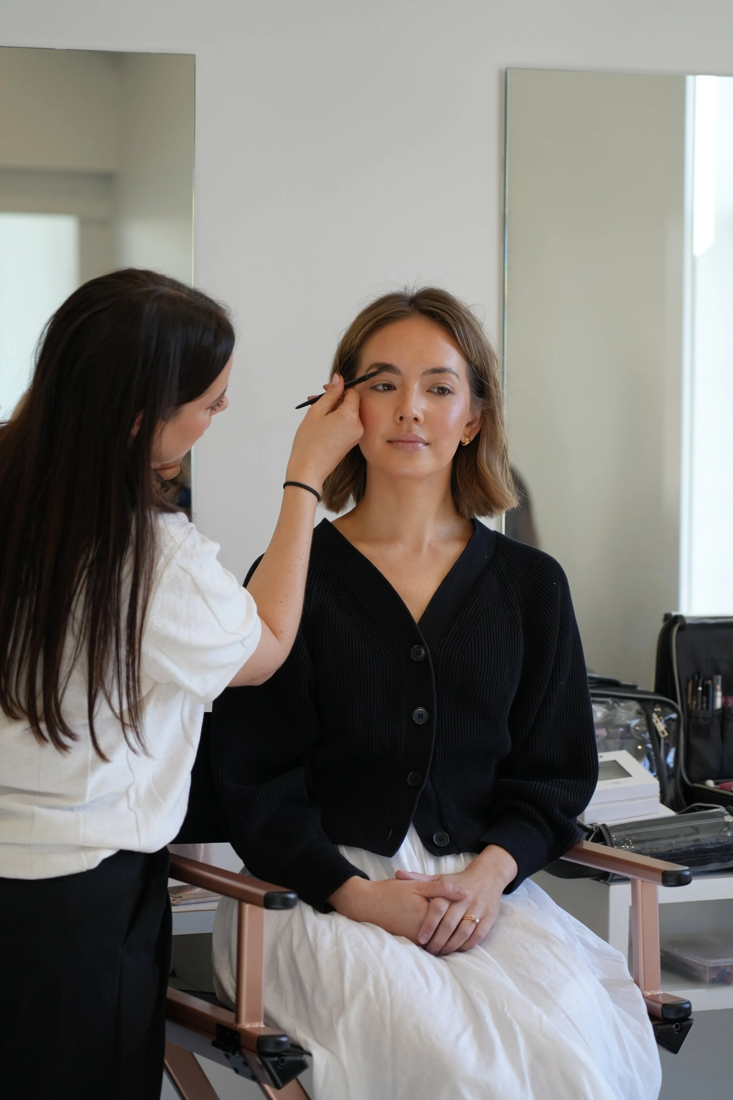 Makeup artist applying eye makeup to a woman seated on a makeup chair in a beauty salon.