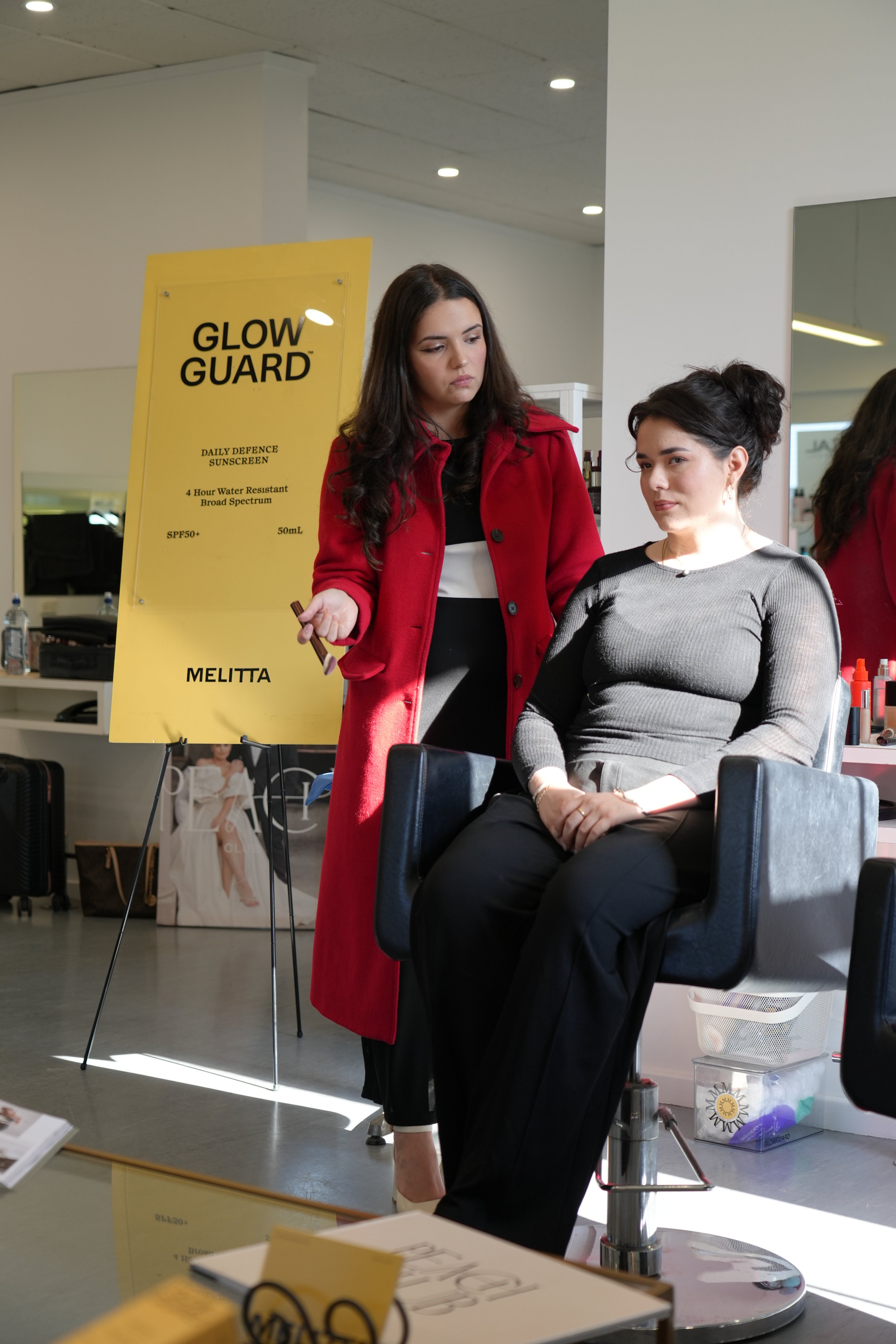 A woman sitting in a salon chair getting her makeup done, with a stylist standing beside her. A sign in the background advertises sunscreen.