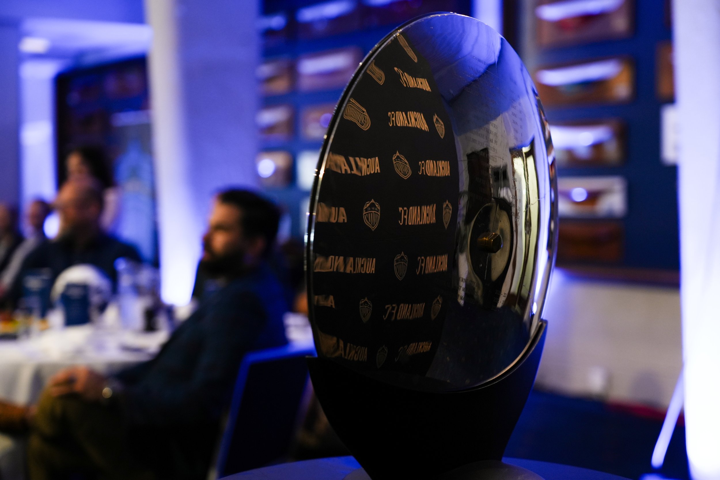 A black, shiny, round object reflecting a room with people and blue lighting, with a logo visible on the surface.
