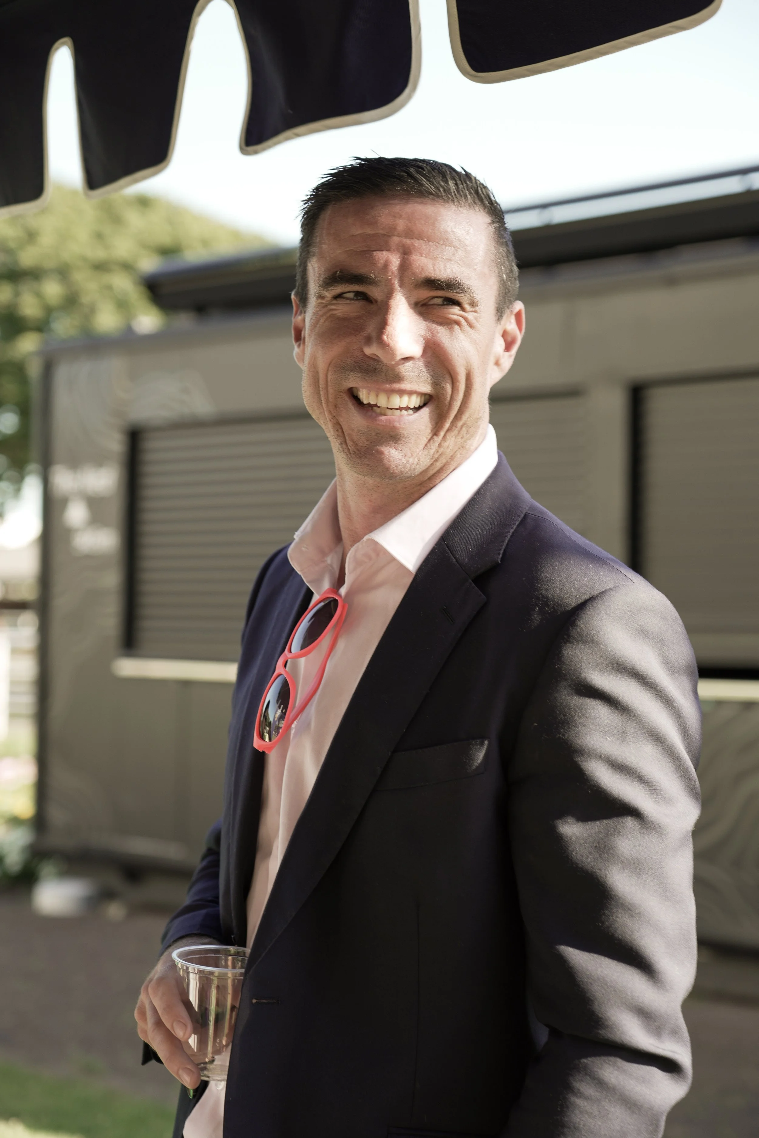 A man dressed in a dark suit with a white shirt, holding a glass of beverage, smiling outdoors with trees and a food truck in the background.