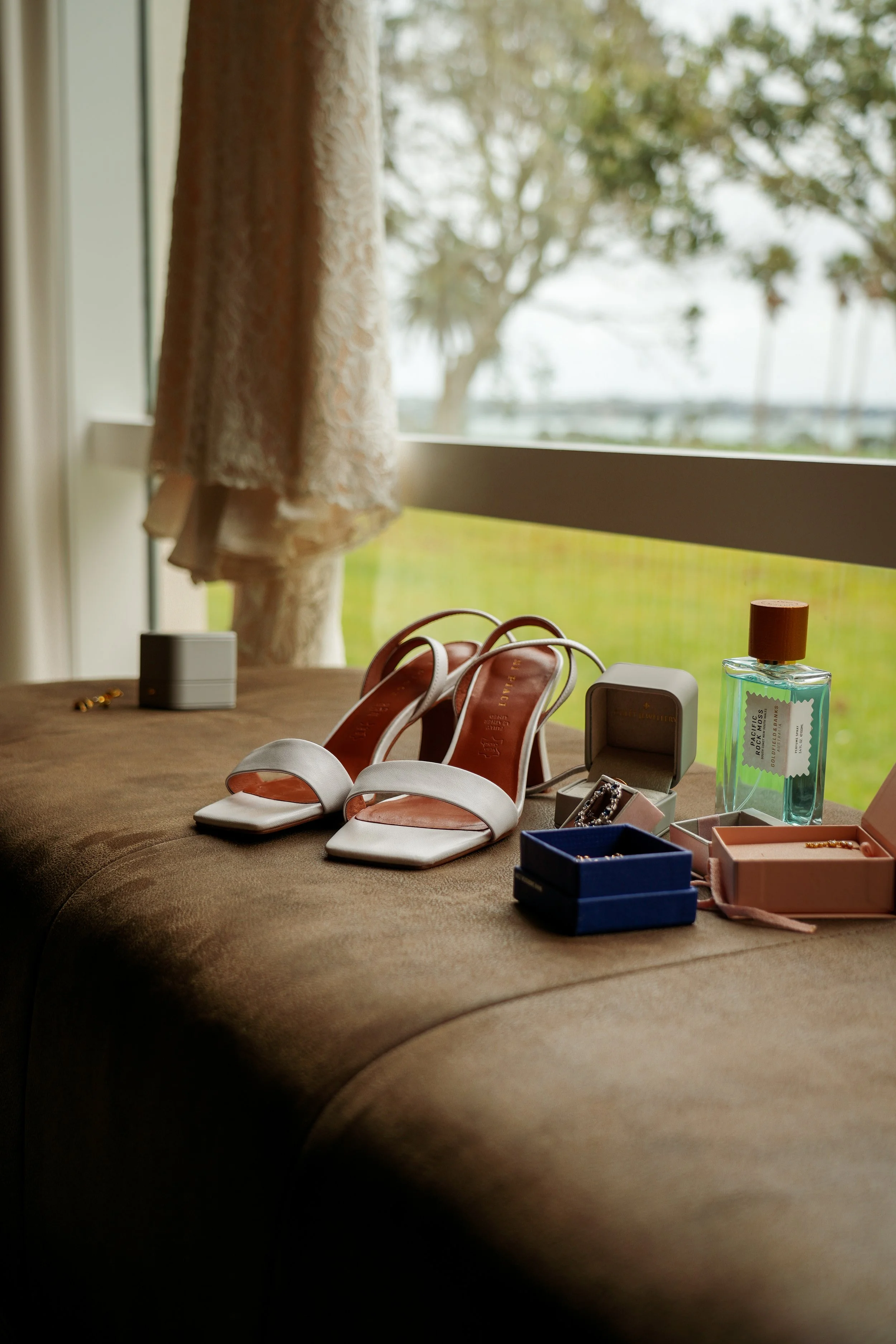 A pair of white high-heeled sandals, jewelry boxes, perfume, and small accessories on a brown surface near a window with green scenery outside.