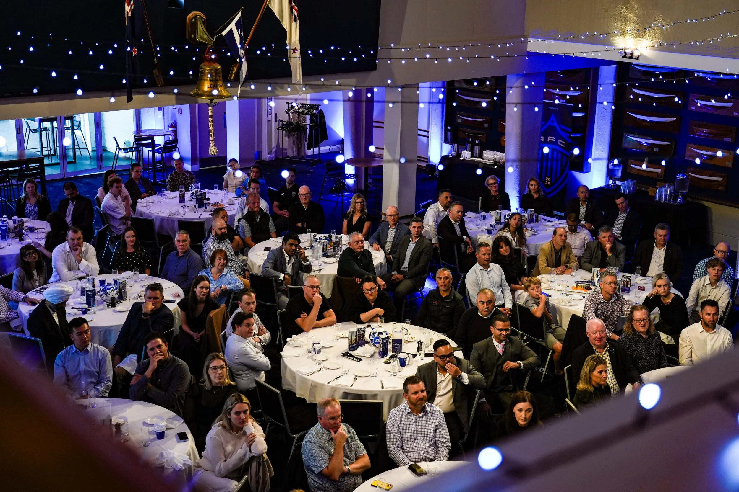 A large group of people seated around multiple round tables at a formal event or dinner, with a decor of string lights and blue accent lighting in the background.