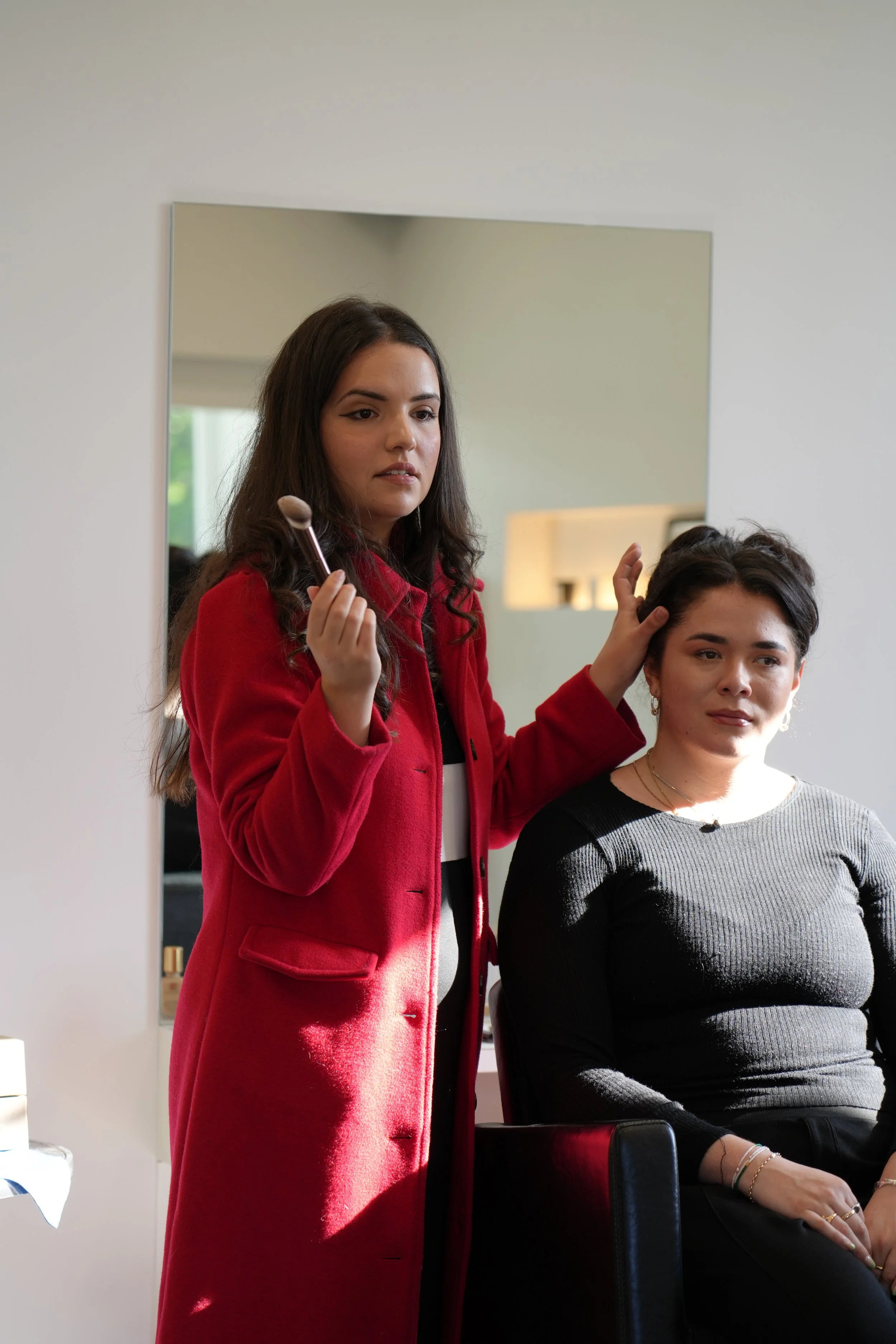 Makeup artist in a red coat applying makeup to a woman sitting in a chair.