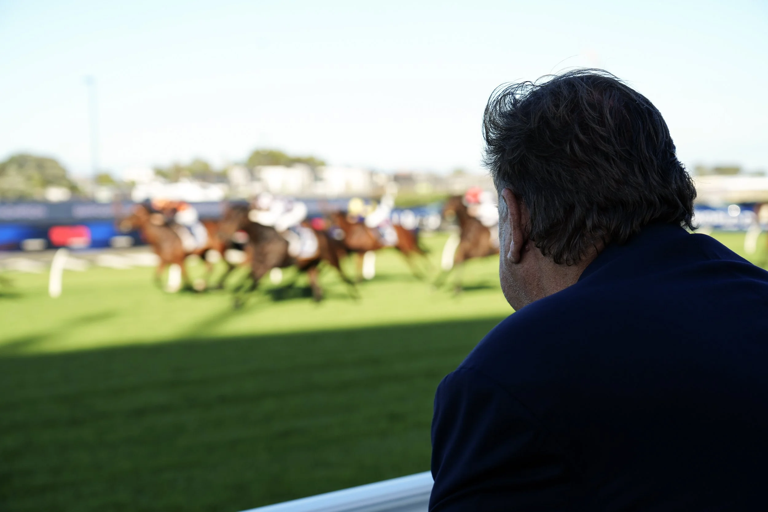 A man watches a horse race from the sidelines with horses and jockeys racing on a green field in the background.