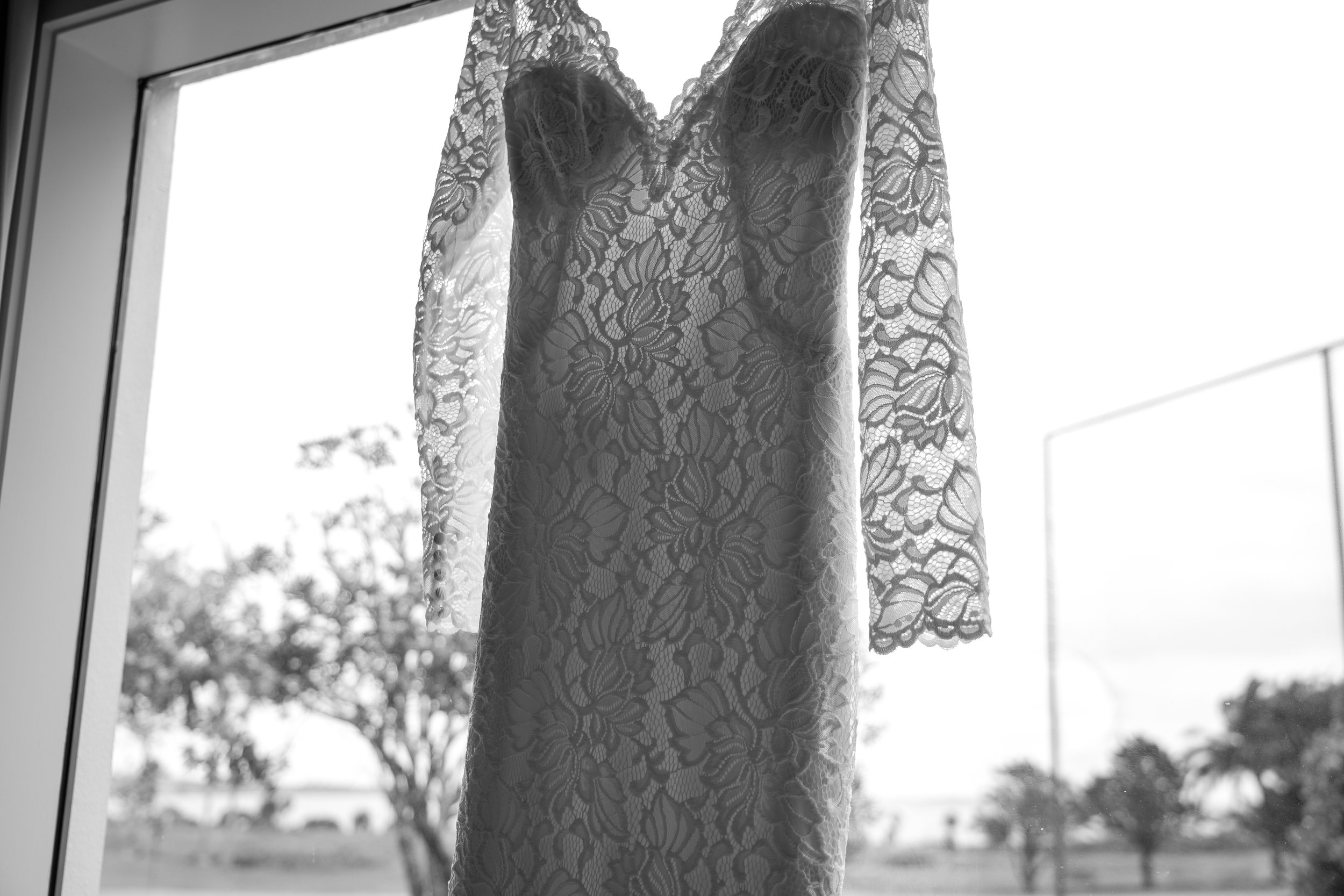 Lace wedding dress hanging in front of a window, with trees and sky visible outside.