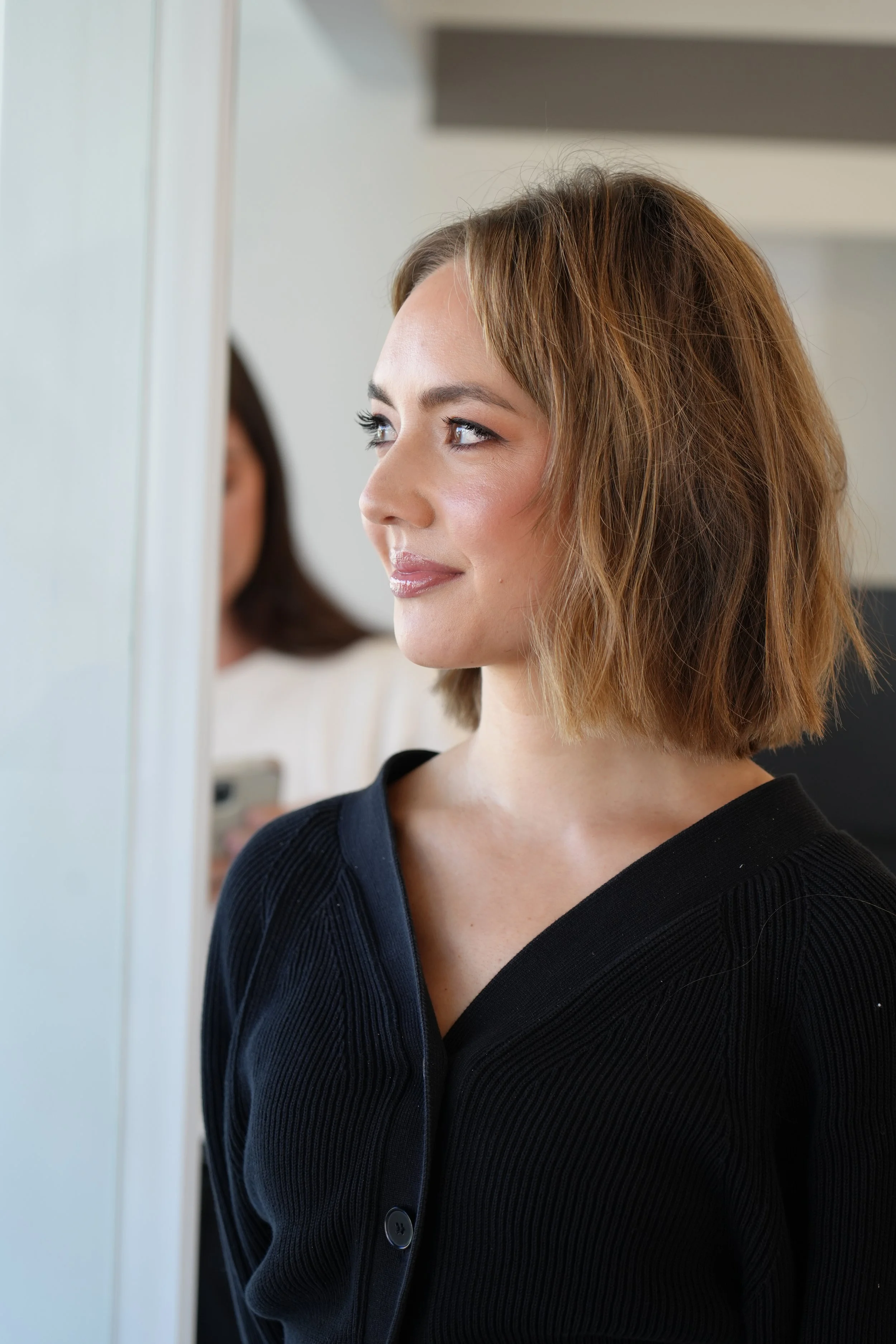 A woman with shoulder-length light brown hair and fair skin is standing in front of a mirror. She is wearing a black, ribbed, button-up top and looking slightly to the side with a slight smile.