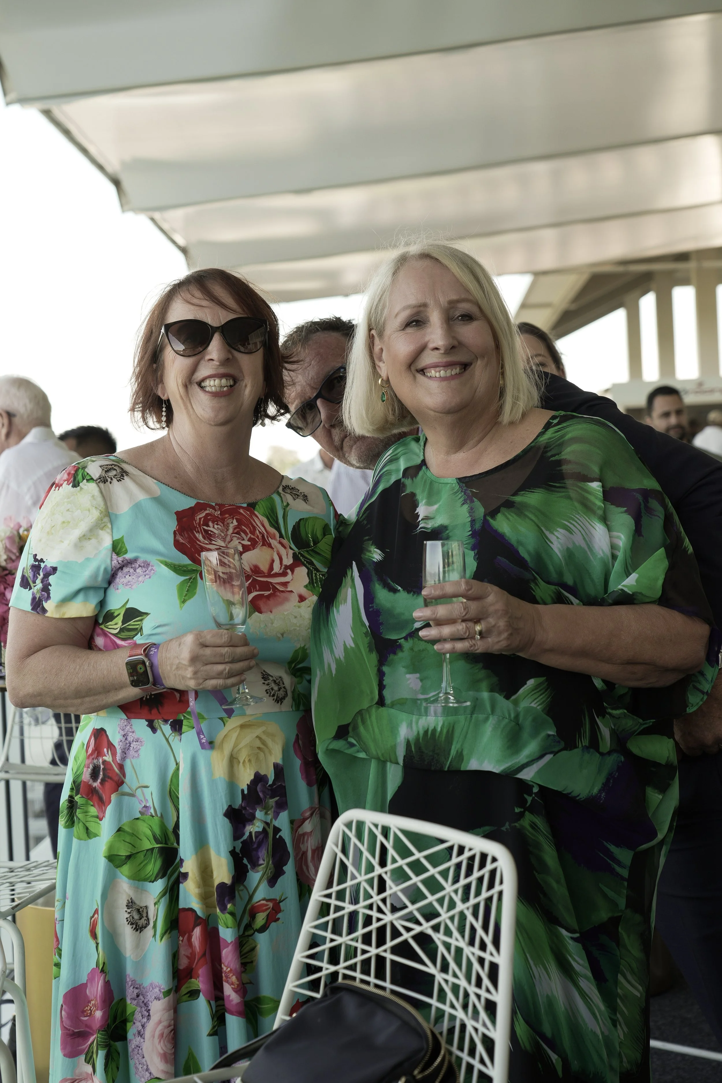 Three people at an outdoor event holding champagne glasses, smiling for the camera. They are dressed in colorful floral clothing, with a woman wearing sunglasses on the left, a woman on the right wearing earrings, and a man partially hidden behind th