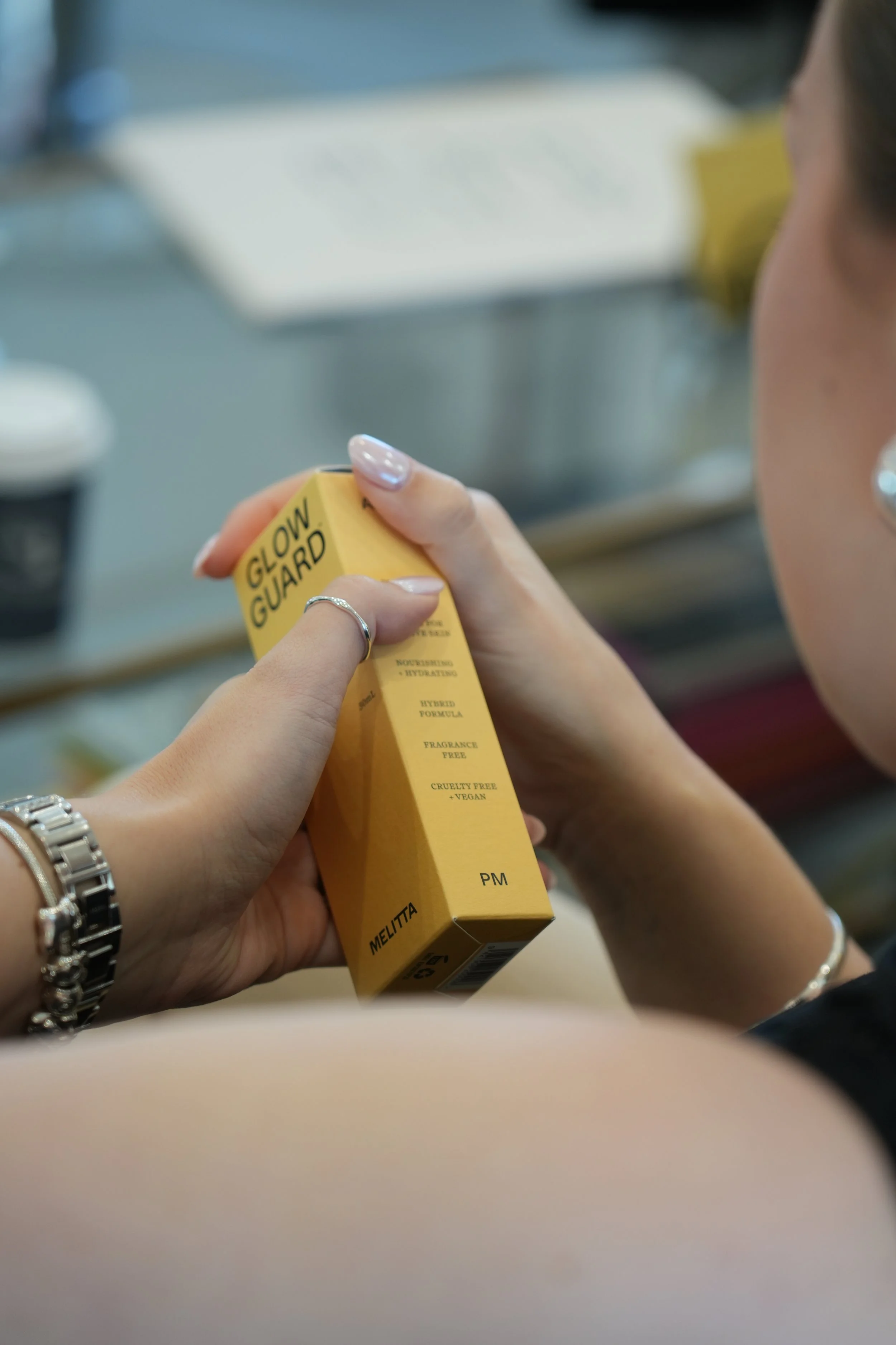 Close-up of a woman holding a yellow box labeled Glow Guard, which is a skincare product, in a retail or pharmacy environment.