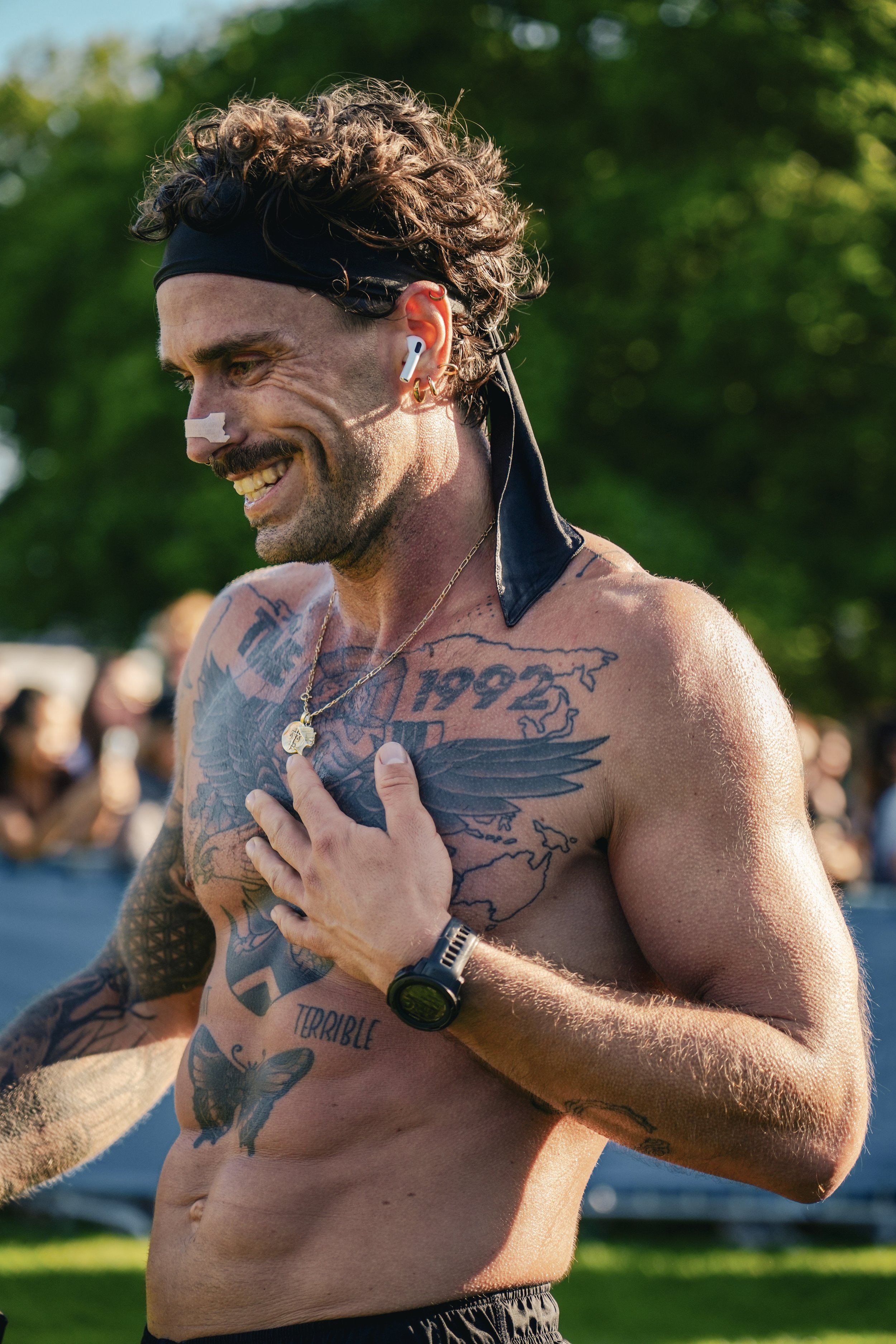 A shirtless man with tattoos and a black headband smiling outdoors, wearing earbuds, a watch, and a gold necklace, with a crowd and green trees in the background.