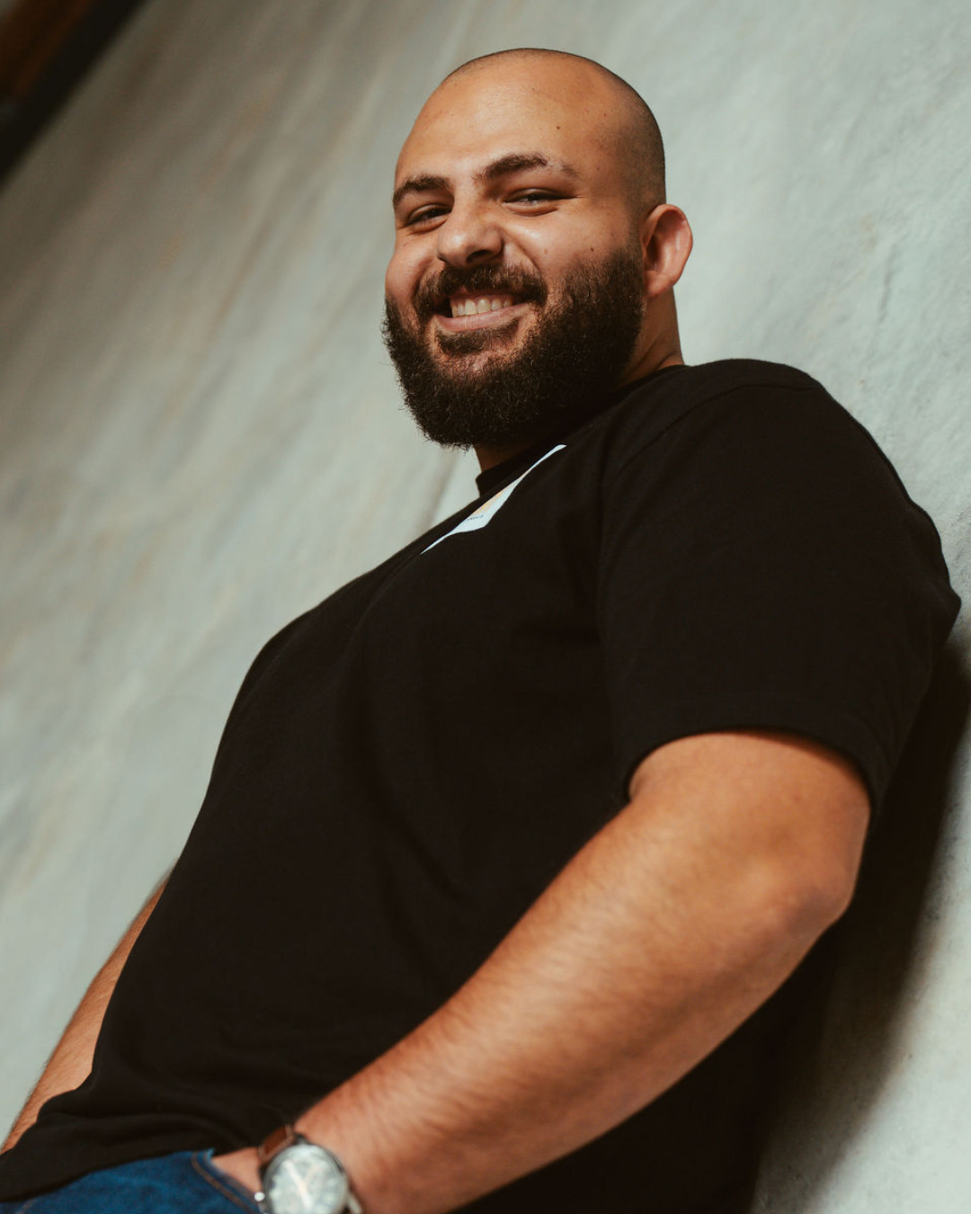A smiling man with a beard and bald head leaning against a wall, wearing a black shirt and a watch.
