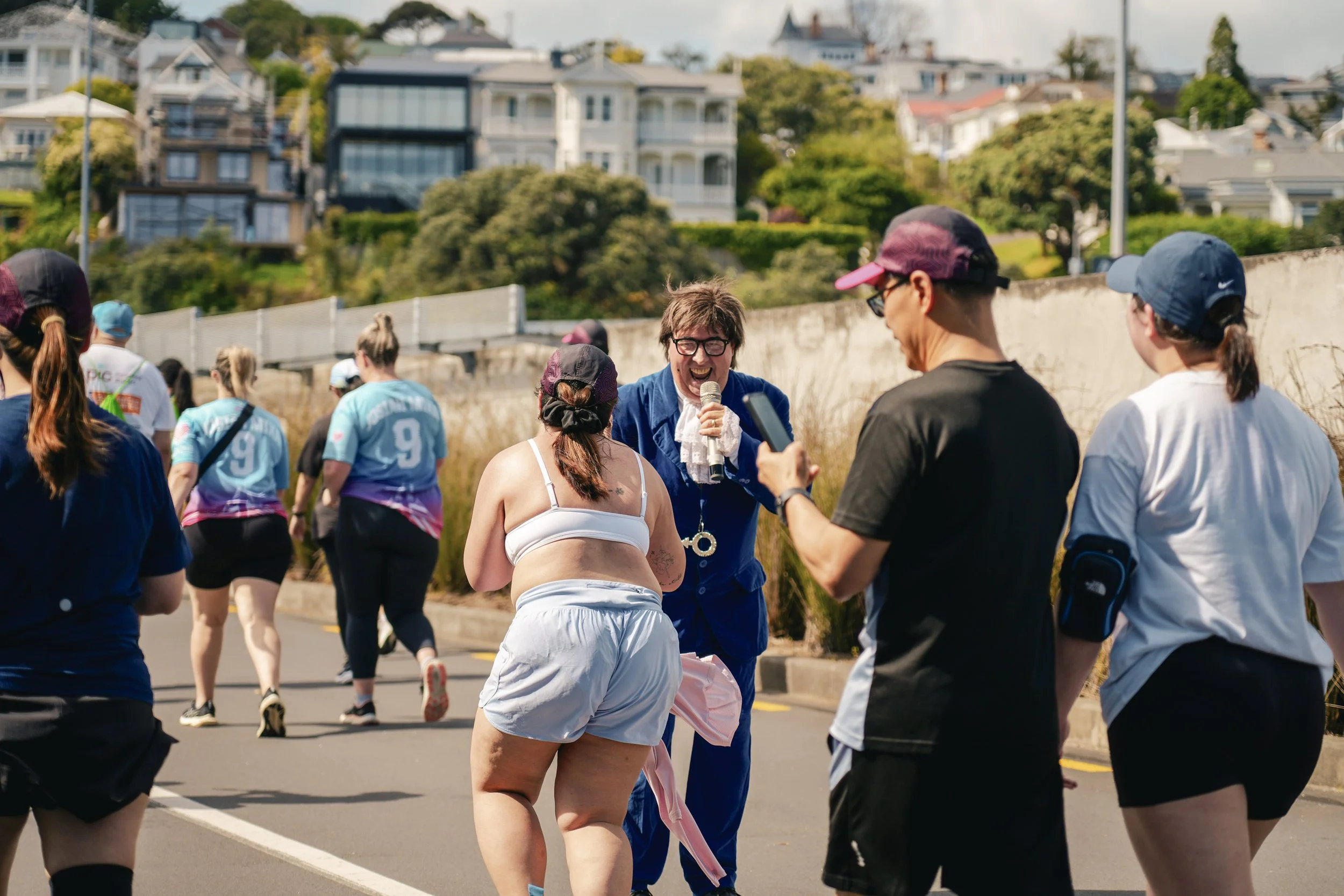 People participating in a marathon or walkathon on a city street, with a man in a blue costume holding a microphone and smiling, as others walk or record with smartphones, and houses and trees in the background.