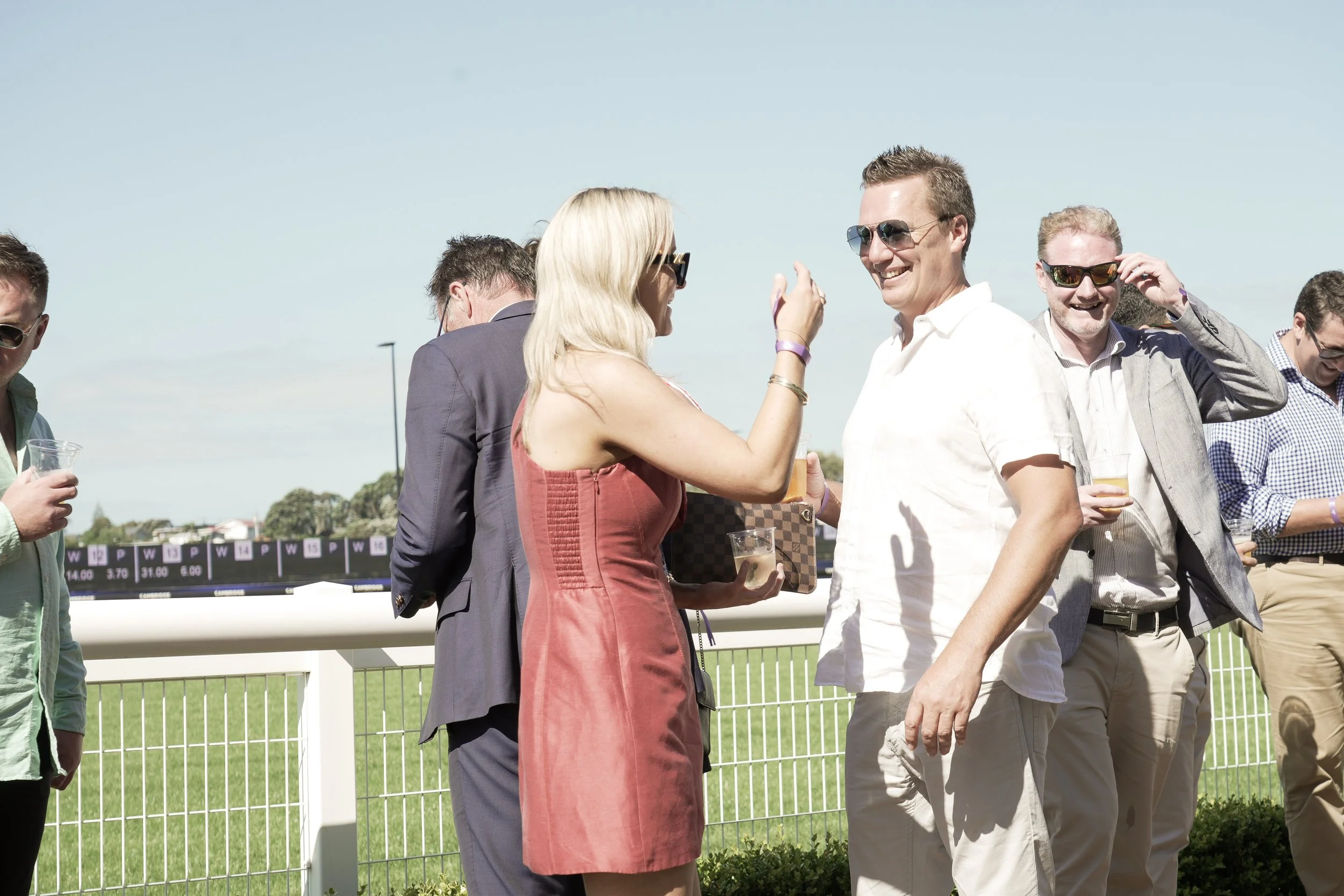 Group of people at an outdoor event, conversing and holding drinks on a sunny day.