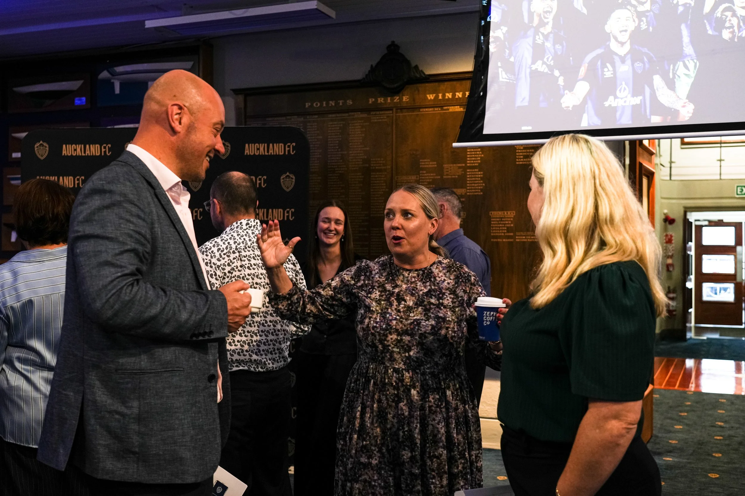 People engaged in conversation at an indoor event with Auckland FC branding and a large screen displaying team images.