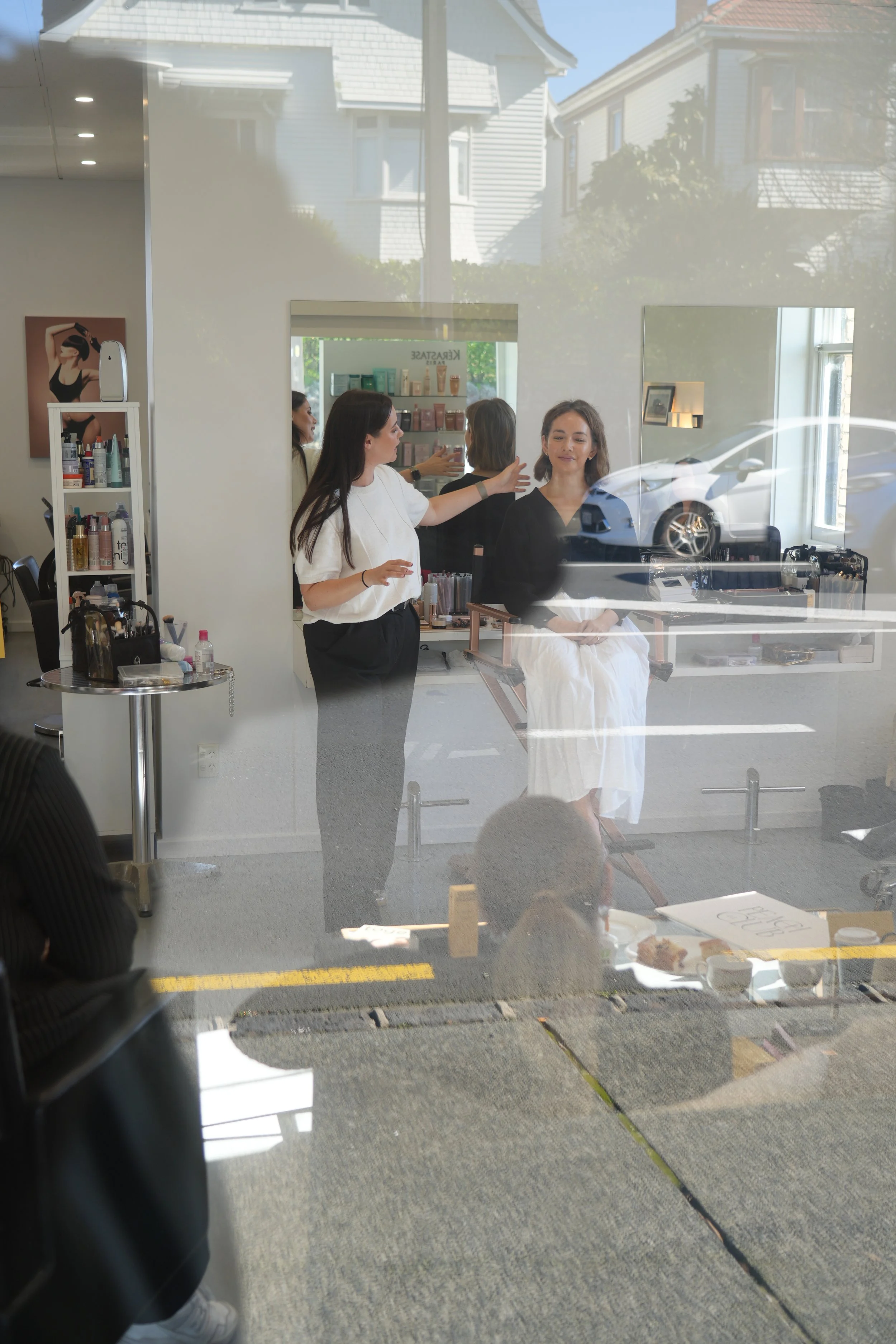 A woman is sitting in a barber chair inside a salon, getting her hair styled by a hairdresser, with various hair products and tools visible around them.