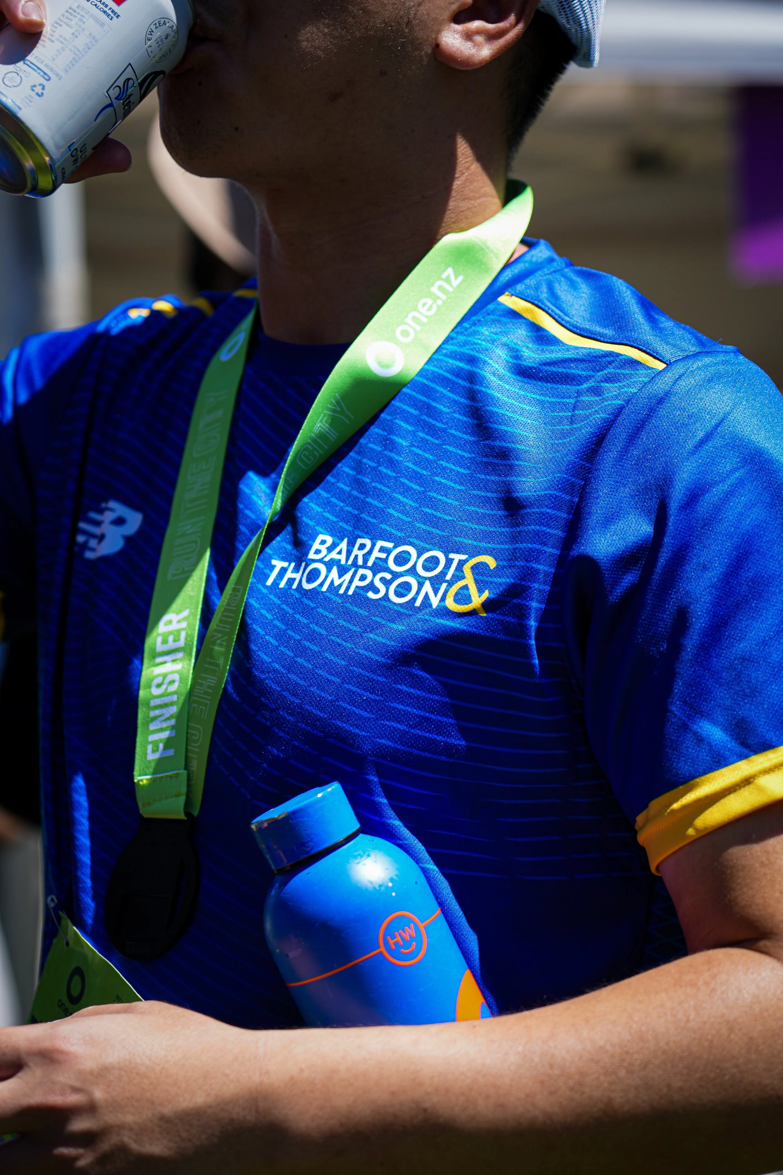 A marathon runner wearing a blue race shirt with yellow accents and a green finisher medal around his neck, drinking from a can and carrying a blue water bottle during a race.