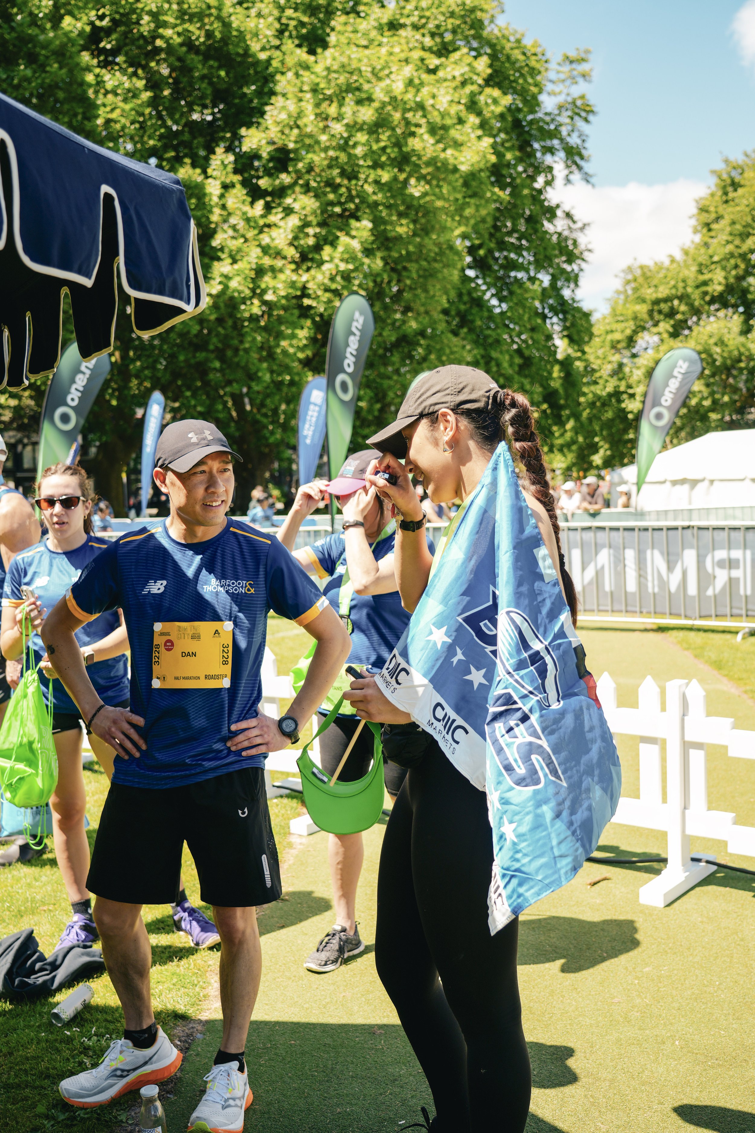 A woman at a marathon event, wearing a dark cap and athletic outfit, is seen smiling as she talks to another woman who is holding a Marathon shirt or flag and has a green Cap and black leggings. Other runners and event banners are visible in the back