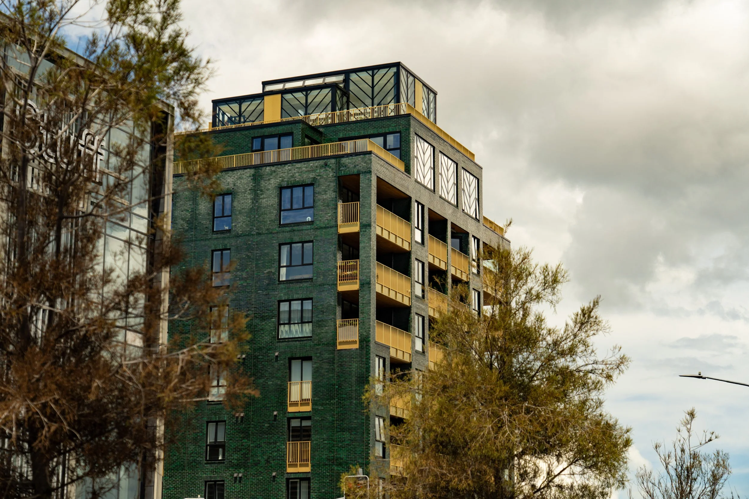 Modern multi-story apartment building with balconies and green brick exterior, partially obscured by trees, under a cloudy sky.
