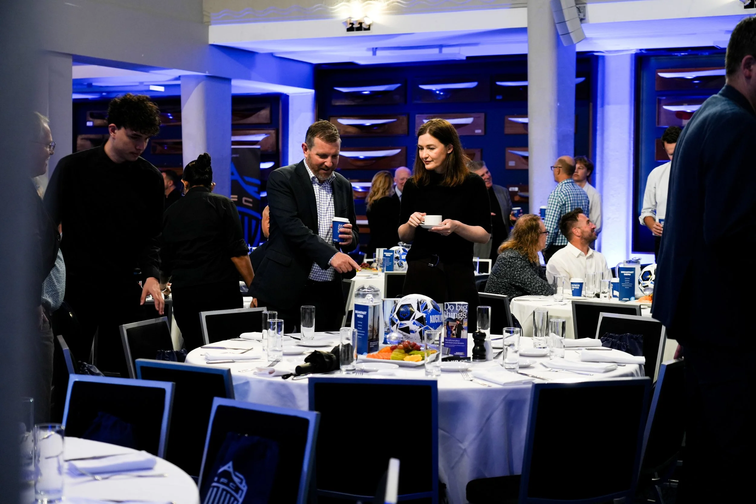 People mingling at a banquet or conference with tables set for dining, decorated with jerseys, fruit, and blue-themed materials, in a room with modern lighting and decor.