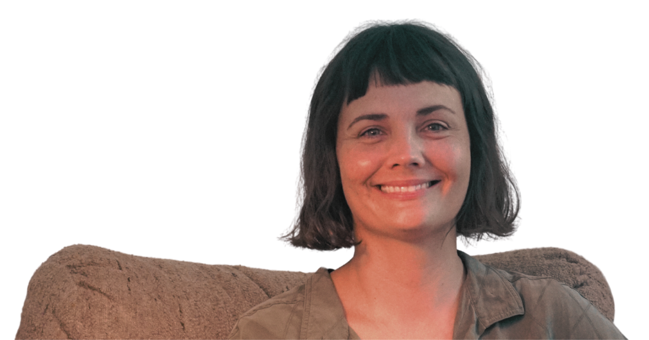 A woman with short dark hair smiling, sitting on a brown couch. She is wearing a khaki shirt and appears to be in a relaxed, casual setting.