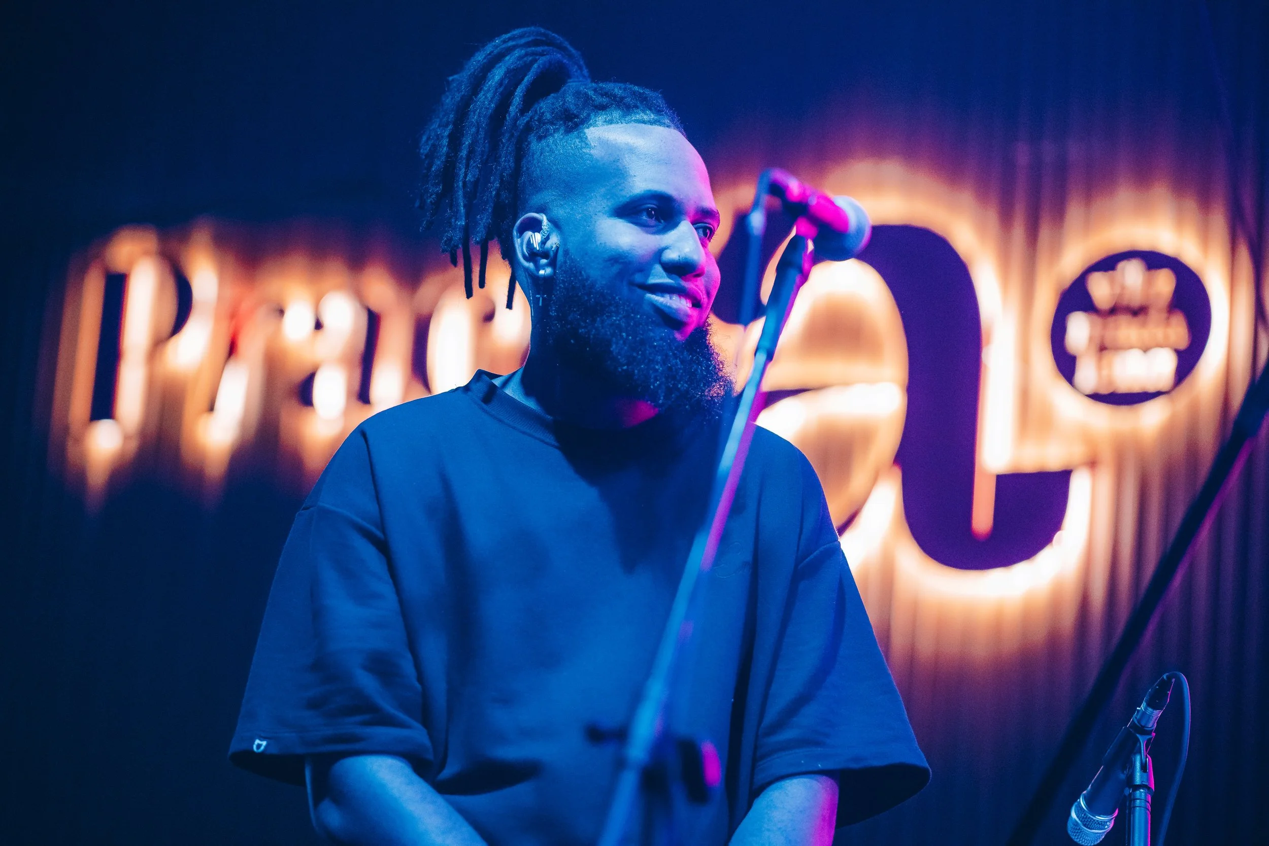 Homem com dreadlocks e barba, sorrindo, de microfone na mão, em palco iluminado com sinal luminoso ao fundo.