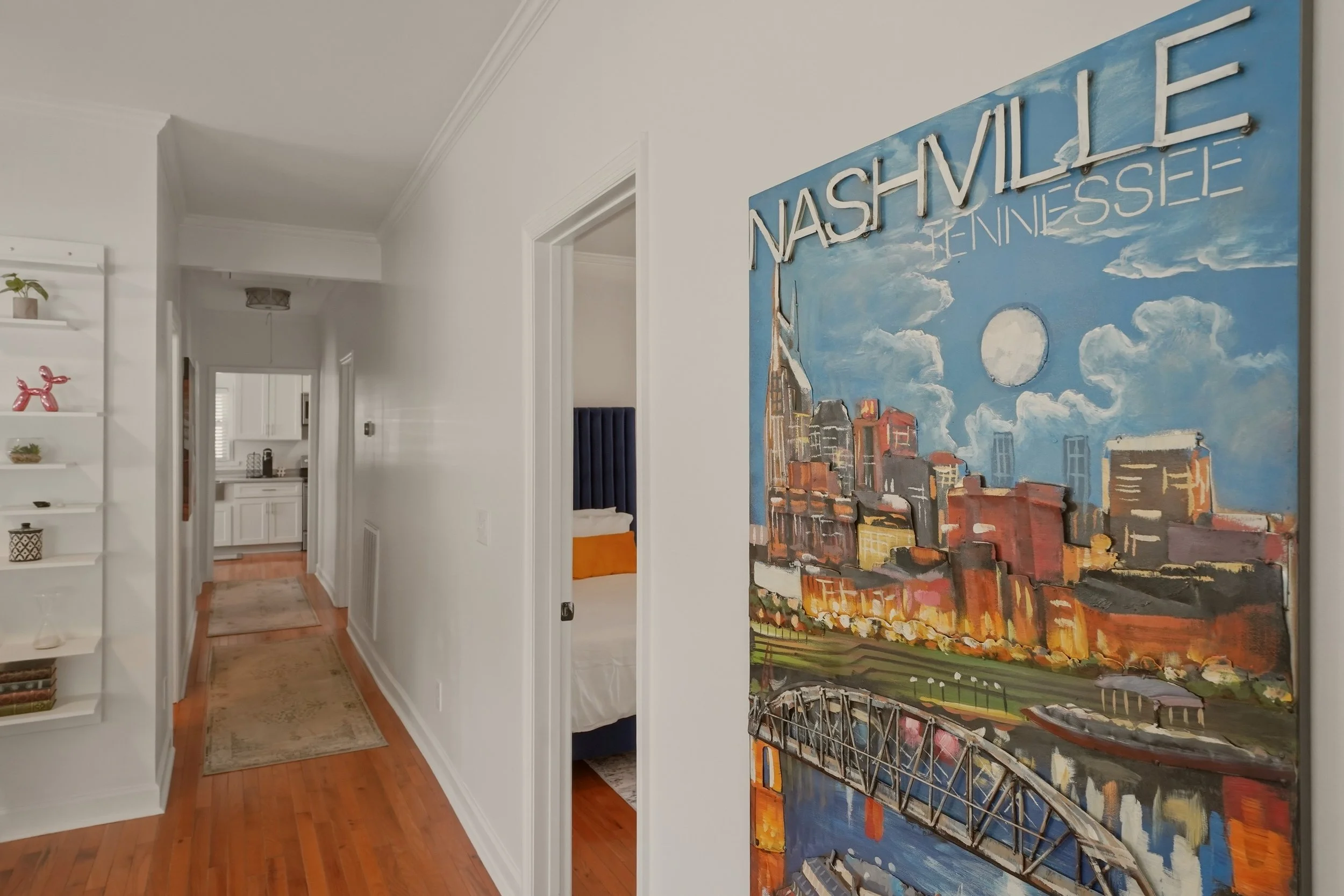 Interior view of a hallway with a white wall, a colorful mural of Nashville, Tennessee, cityscape, and a large blue sky with clouds, leading to a partially visible kitchen and bedroom.