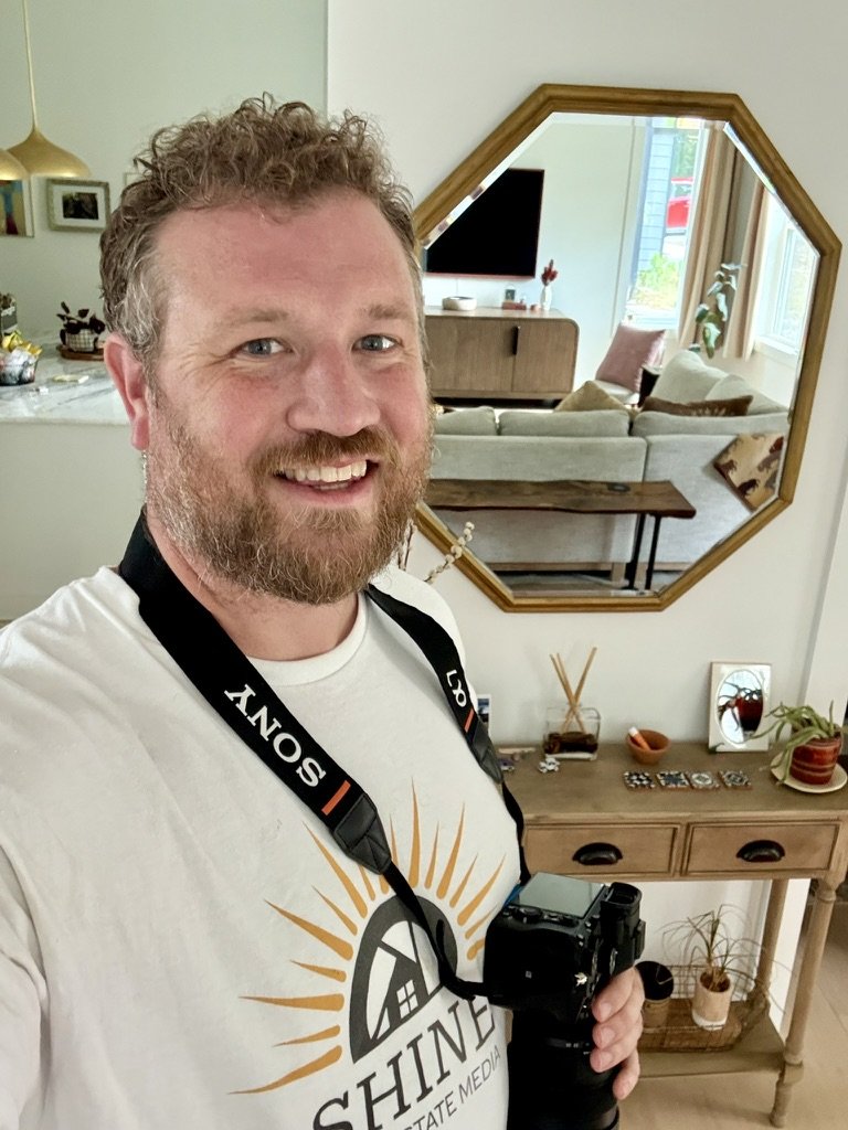A man taking a selfie in a room with a camera hanging around his neck. He has curly hair and a beard, smiling at the camera. The background shows a mirror reflecting a living room with a couch, cushions, and a coffee table, along with some decor items on a wooden console.