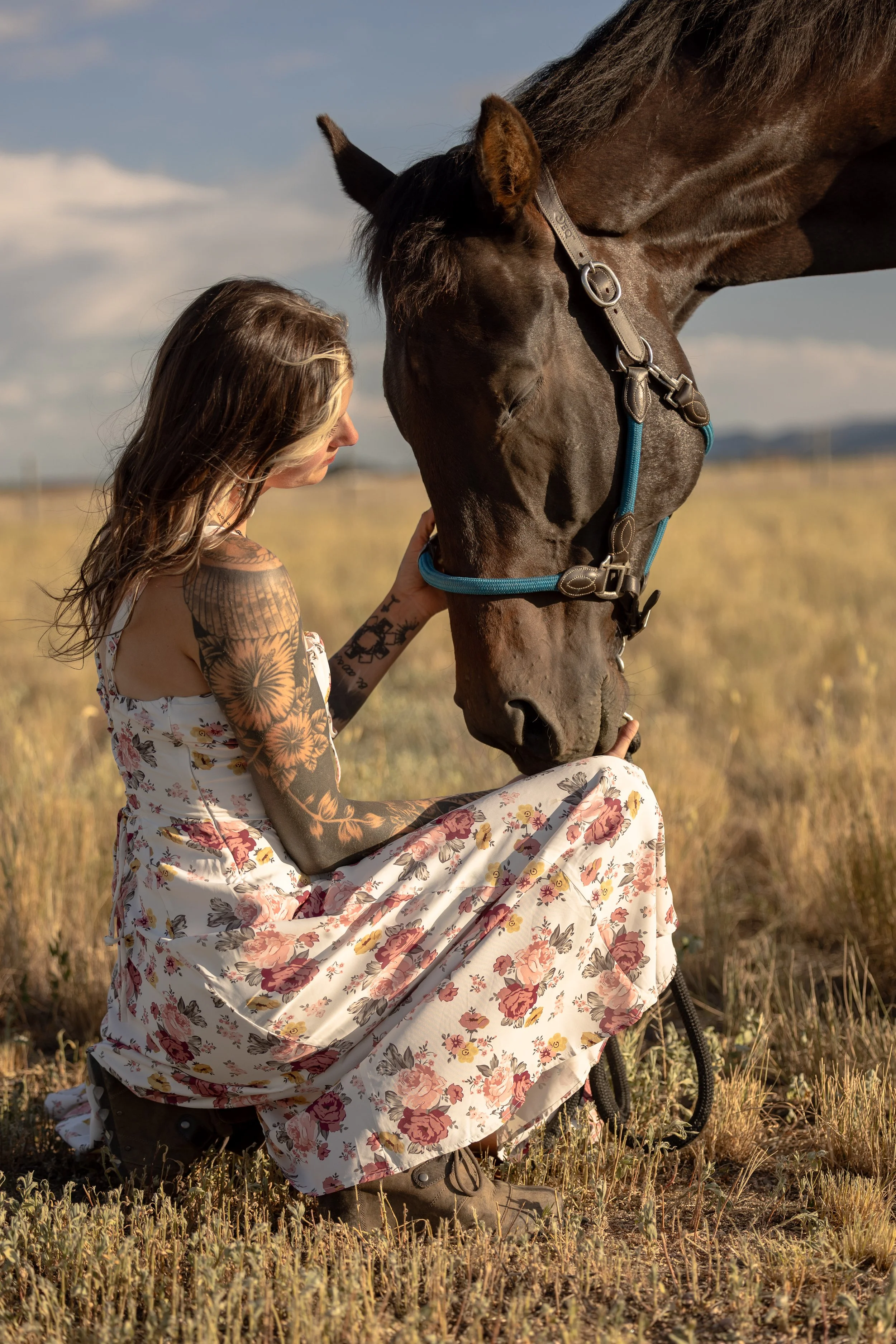 A woman with tattoos kneels in a field to gently hold a large dark brown horse's face, in a peaceful outdoor setting with clear skies.
