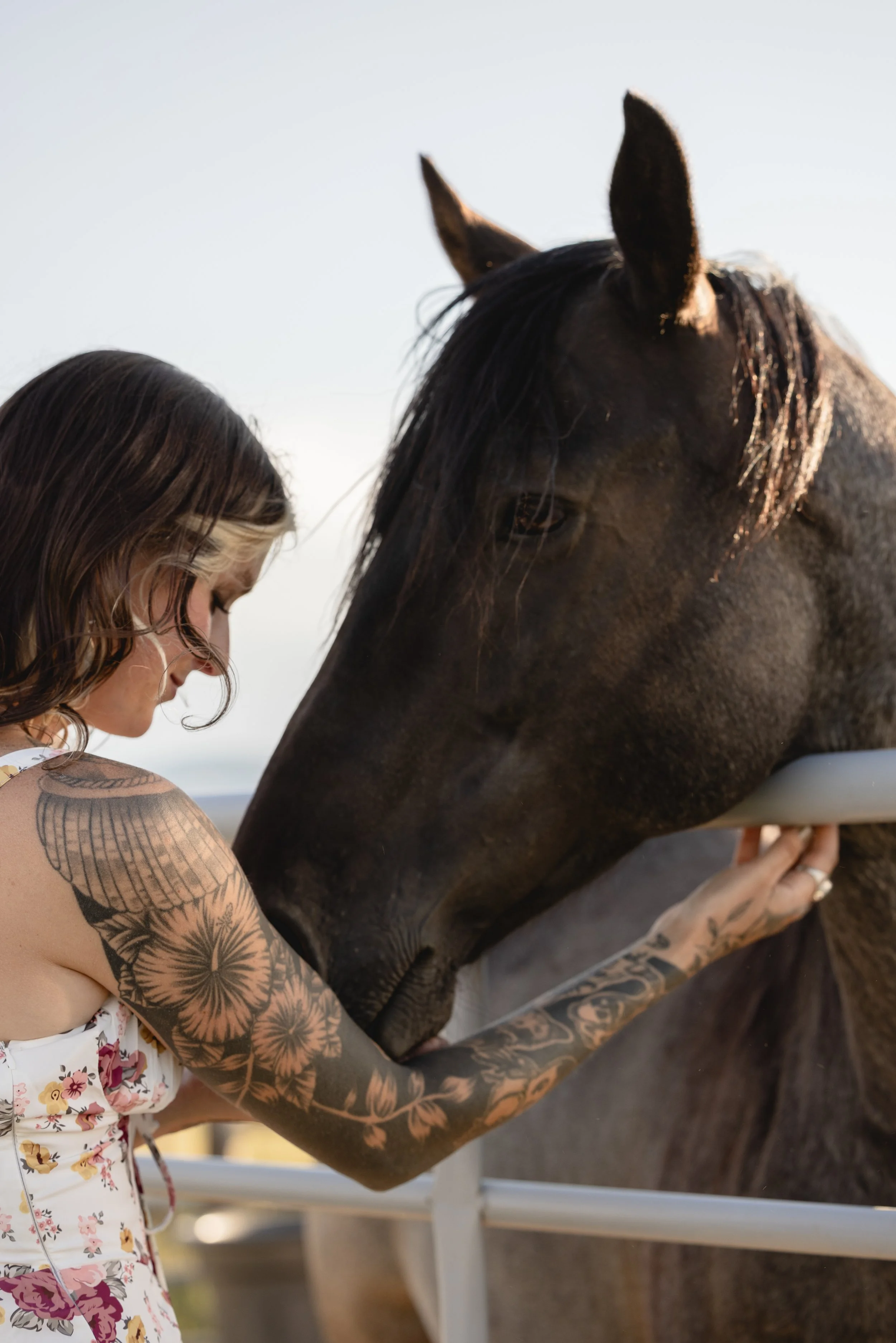 A woman with floral tattoo sleeves is gently petting a dark-colored horse.