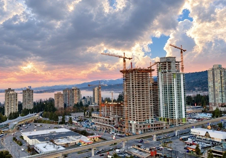 City skyline at sunset with skyscrapers, including some under construction with cranes, and a populated parking lot below.