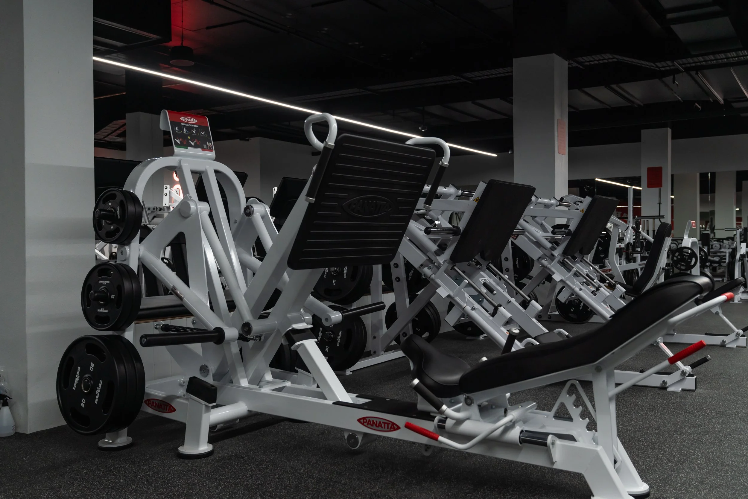 Various gym equipment including leg press and weight machines in an indoor fitness center with dark ceiling and bright lighting.