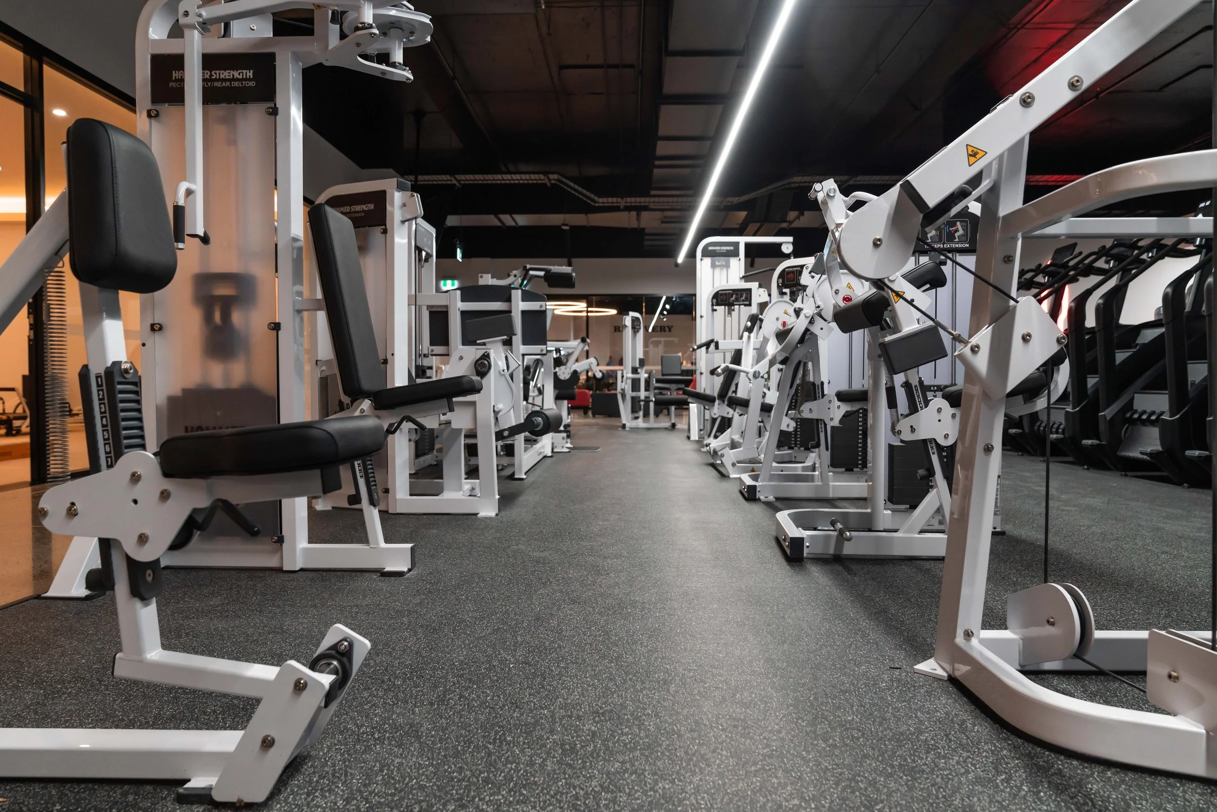 gym with various white and black exercise machines lined up on a dark floor, illuminated by overhead lighting.